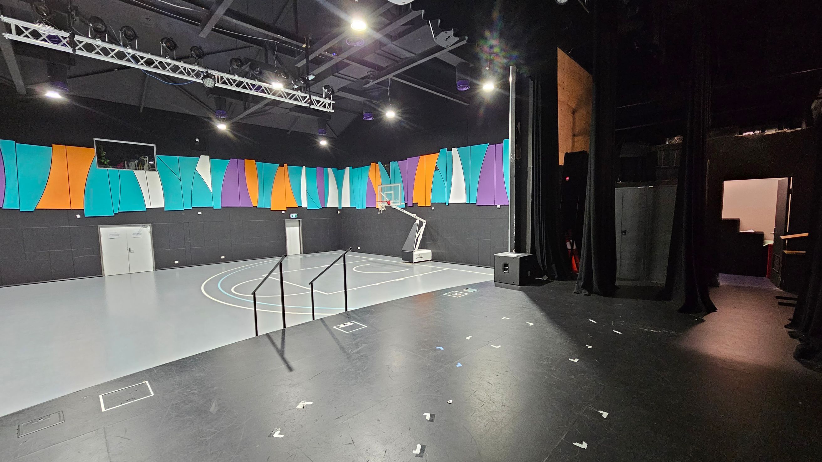 A multi-purpose indoor space with a basketball court on the left, featuring a hoop and court markings. The walls are adorned with colourful panels in orange, teal, purple, and white. The ceiling has exposed beams and lighting fixtures. On the right, there is a stage area with black curtains and a speaker. A doorway leads to a backstage area. The floor is a mix of grey and black surfaces. A multi-purpose indoor space with a basketball court on the left, featuring a hoop and court markings. The walls are adorned with colourful panels in orange, teal, purple, and white. The ceiling has exposed beams and lighting fixtures. On the right, there is a stage area with black curtains and a speaker. A doorway leads to a backstage area. The floor is a mix of grey and black surfaces.