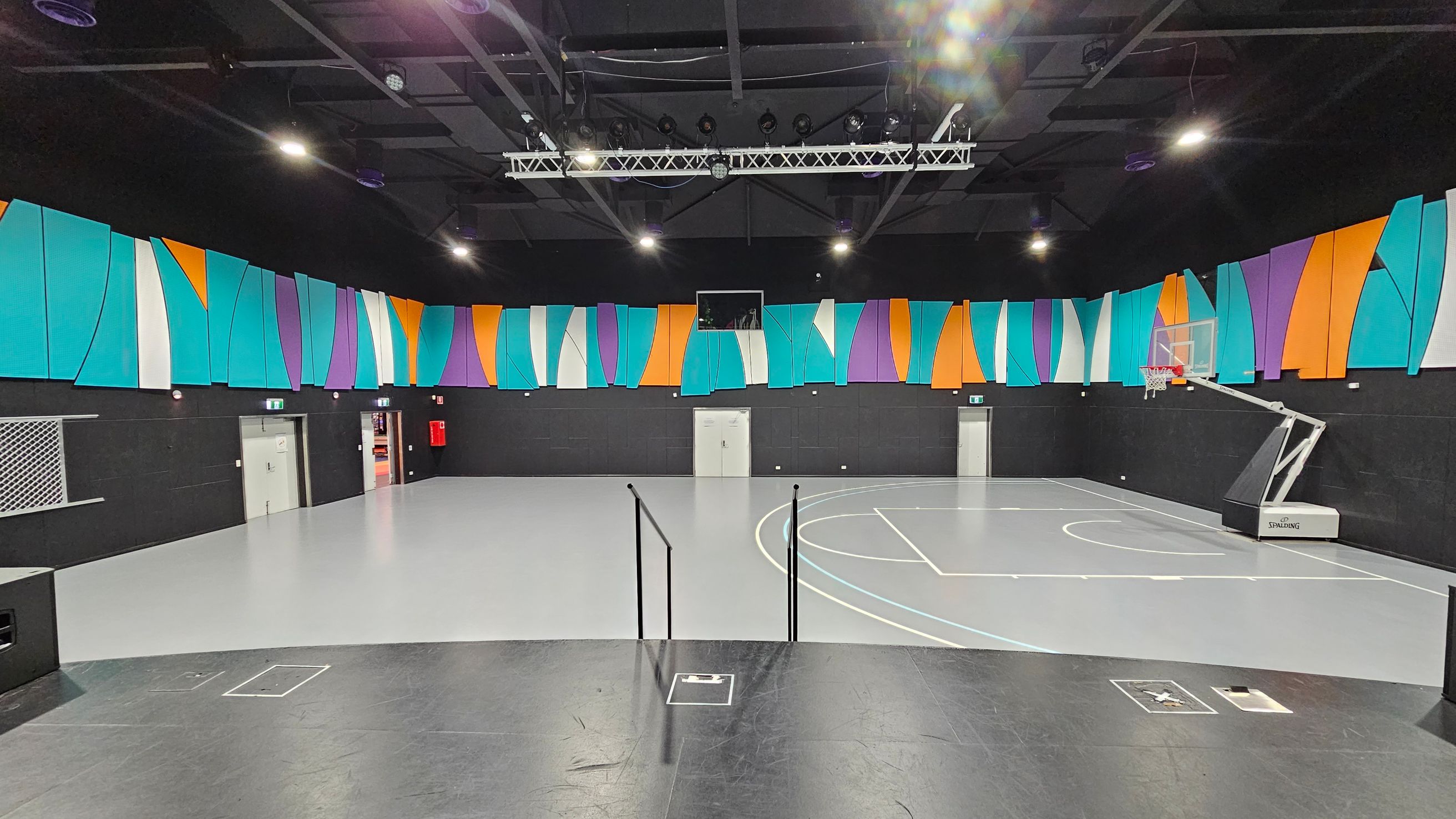 A modern indoor basketball court with a grey floor and a black ceiling. The walls are adorned with colourful, abstract panels in shades of turquoise, orange, purple, and white. A basketball hoop is positioned on the right side. There are multiple doors along the back wall, and a fire extinguisher is visible. Overhead, there are lights and a metal truss system. The foreground features a black platform with steps leading down to the court. A modern indoor basketball court with a grey floor and a black ceiling. The walls are adorned with colourful, abstract panels in shades of turquoise, orange, purple, and white. A basketball hoop is positioned on the right side. There are multiple doors along the back wall, and a fire extinguisher is visible. Overhead, there are lights and a metal truss system. The foreground features a black platform with steps leading down to the court.