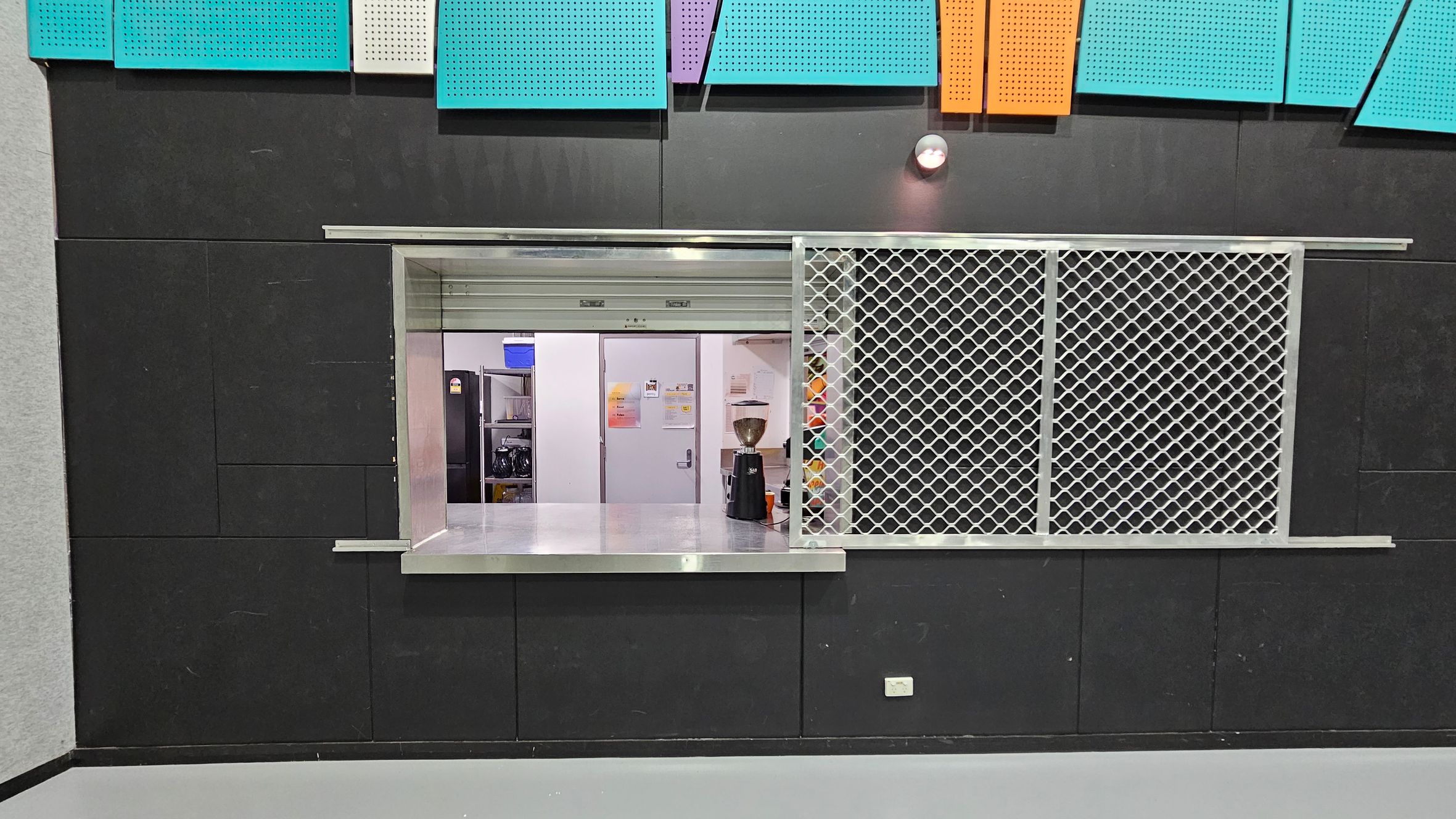 A service counter with a metal mesh screen on the right and an open window on the left. Behind the counter, there is a white door and shelves with various items, including a blue container and a coffee grinder. The wall is black with colourful acoustic panels in teal, purple, and orange at the top. The floor is grey, and there is a small light fixture above the counter. A service counter with a metal mesh screen on the right and an open window on the left. Behind the counter, there is a white door and shelves with various items, including a blue container and a coffee grinder. The wall is black with colourful acoustic panels in teal, purple, and orange at the top. The floor is grey, and there is a small light fixture above the counter.