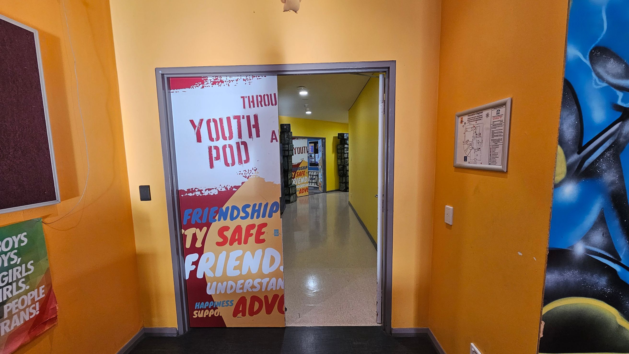 A hallway with bright yellow walls. On the left, a partially visible poster with colourful text, including "YOUTH POD" and words like "FRIENDSHIP" and "SAFE". A bulletin board is also on the left. On the right, a framed map or notice is mounted on the wall, and part of a blue and black mural is visible. The floor is shiny and light-coloured, leading to another room with similar decor. Ceiling lights illuminate the space. A hallway with bright yellow walls. On the left, a partially visible poster with colourful text, including "YOUTH POD" and words like "FRIENDSHIP" and "SAFE". A bulletin board is also on the left. On the right, a framed map or notice is mounted on the wall, and part of a blue and black mural is visible. The floor is shiny and light-coloured, leading to another room with similar decor. Ceiling lights illuminate the space.