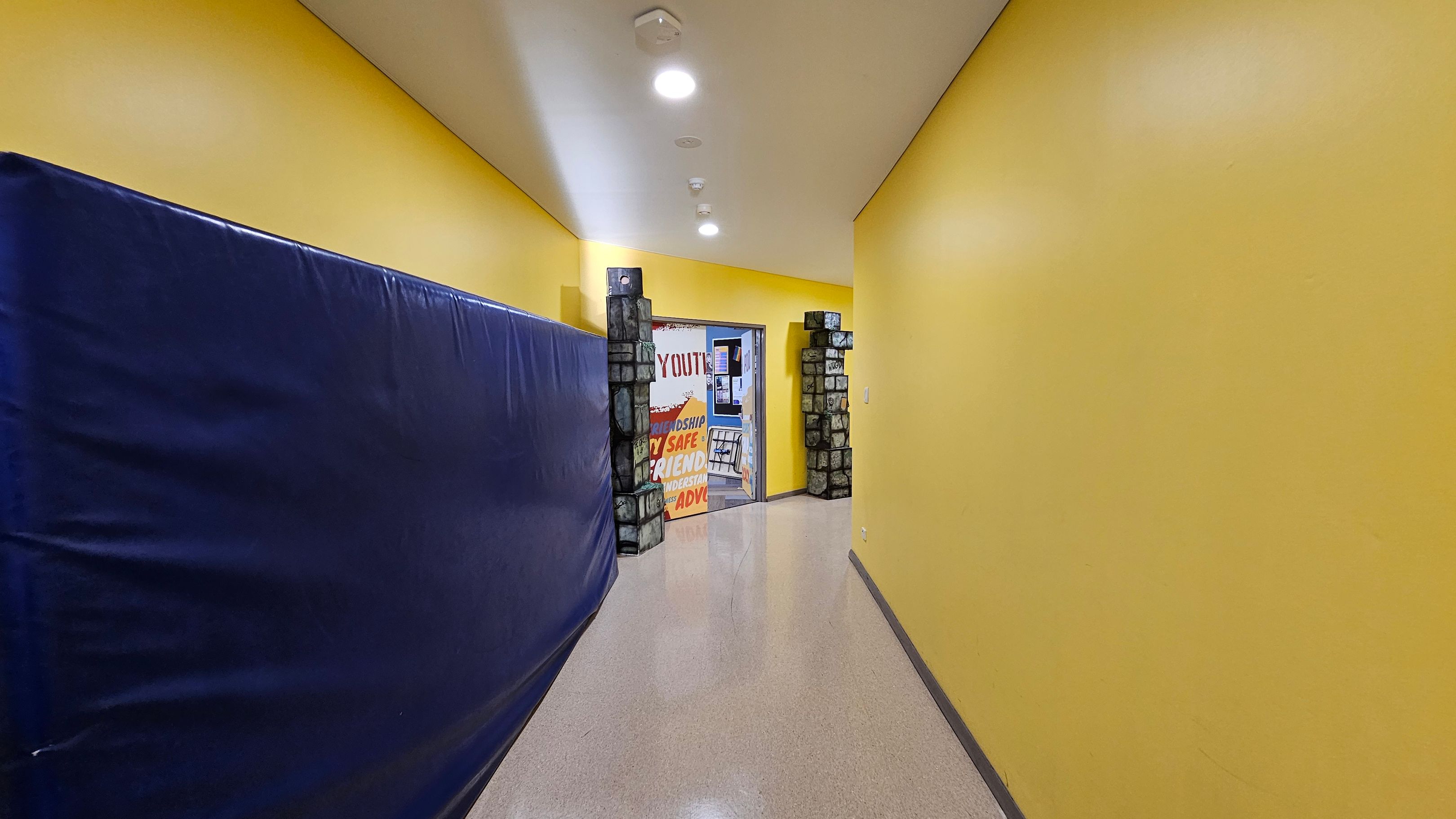 A hallway with bright yellow walls and a glossy floor. On the left, a large blue padded mat covers the wall. At the end of the hallway, there's an open doorway framed by decorative stone-like pillars. Through the doorway, a colourful sign with words like "YOUTH," "SAFE," and "FRIENDSHIP" is visible. The ceiling is white with recessed lighting. A hallway with bright yellow walls and a glossy floor. On the left, a large blue padded mat covers the wall. At the end of the hallway, there's an open doorway framed by decorative stone-like pillars. Through the doorway, a colourful sign with words like "YOUTH," "SAFE," and "FRIENDSHIP" is visible. The ceiling is white with recessed lighting.