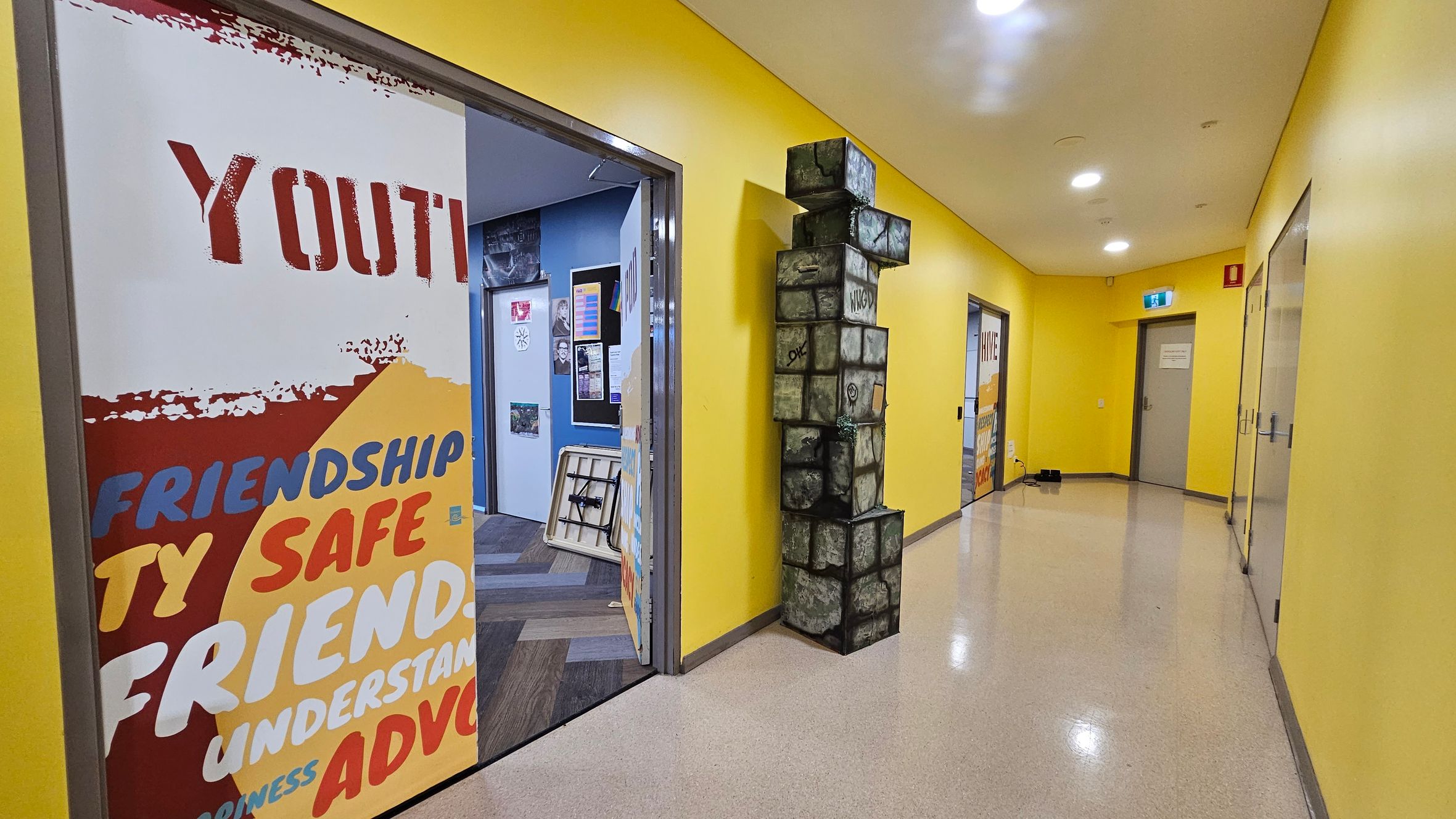 A hallway with bright yellow walls and a shiny, light-coloured floor. On the left, a door is open, revealing a room with blue walls and various posters. A large poster on the door displays words like "YOUTH," "FRIENDSHIP," and "SAFE." In the middle, a stack of cube-shaped blocks with a stone-like appearance is positioned against the wall. On the right, another door is visible, and further down the hallway, an exit sign is mounted above a closed door. Ceiling lights illuminate the space. A hallway with bright yellow walls and a shiny, light-coloured floor. On the left, a door is open, revealing a room with blue walls and various posters. A large poster on the door displays words like "YOUTH," "FRIENDSHIP," and "SAFE." In the middle, a stack of cube-shaped blocks with a stone-like appearance is positioned against the wall. On the right, another door is visible, and further down the hallway, an exit sign is mounted above a closed door. Ceiling lights illuminate the space.