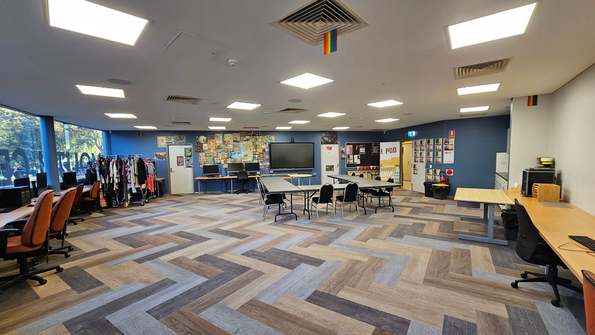 A spacious room with a herringbone-patterned floor in various shades of brown and grey. On the left, there are several orange office chairs in front of computer desks, with a clothing rack nearby. The centre features a large screen on the wall, surrounded by a collage of posters. In the middle, tables and chairs are arranged. On the right, more desks and chairs are visible. The ceiling has bright square lights and a rainbow flag. The walls are blue and white, with more posters and a door. A spacious room with a herringbone-patterned floor in various shades of brown and grey. On the left, there are several orange office chairs in front of computer desks, with a clothing rack nearby. The centre features a large screen on the wall, surrounded by a collage of posters. In the middle, tables and chairs are arranged. On the right, more desks and chairs are visible. The ceiling has bright square lights and a rainbow flag. The walls are blue and white, with more posters and a door.