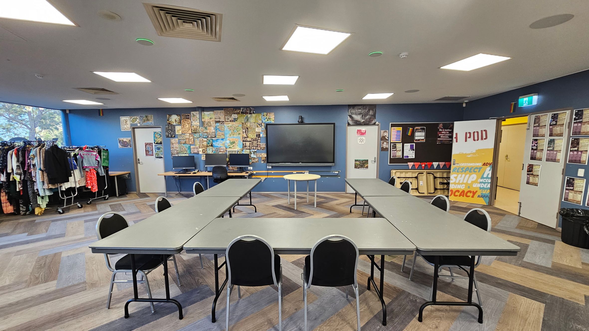 A room with a U-shaped arrangement of tables and chairs in the centre. The floor has a multicoloured wood pattern. The back wall is blue, featuring a large display screen flanked by two computers on a desk. Posters and maps cover the wall. On the left, clothing racks are filled with various garments. On the right, a door with a "POD" sign and a bulletin board with notices. The ceiling has square lights and air vents. A room with a U-shaped arrangement of tables and chairs in the centre. The floor has a multicoloured wood pattern. The back wall is blue, featuring a large display screen flanked by two computers on a desk. Posters and maps cover the wall. On the left, clothing racks are filled with various garments. On the right, a door with a "POD" sign and a bulletin board with notices. The ceiling has square lights and air vents.