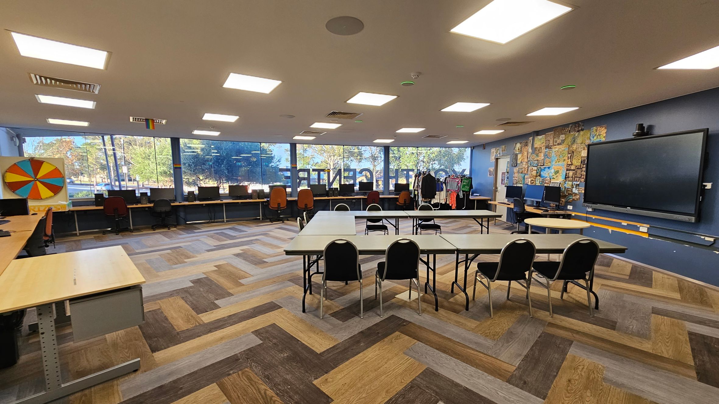 A spacious room with a herringbone-patterned wooden floor. On the left, a colourful wheel is mounted on the wall above a row of desks with computers and chairs. The back wall features large windows with a view of trees outside. In the centre, several tables are arranged in a U-shape with chairs around them. On the right, a large screen is mounted on a wall decorated with various posters and images. The ceiling has multiple rectangular lights. A spacious room with a herringbone-patterned wooden floor. On the left, a colourful wheel is mounted on the wall above a row of desks with computers and chairs. The back wall features large windows with a view of trees outside. In the centre, several tables are arranged in a U-shape with chairs around them. On the right, a large screen is mounted on a wall decorated with various posters and images. The ceiling has multiple rectangular lights.