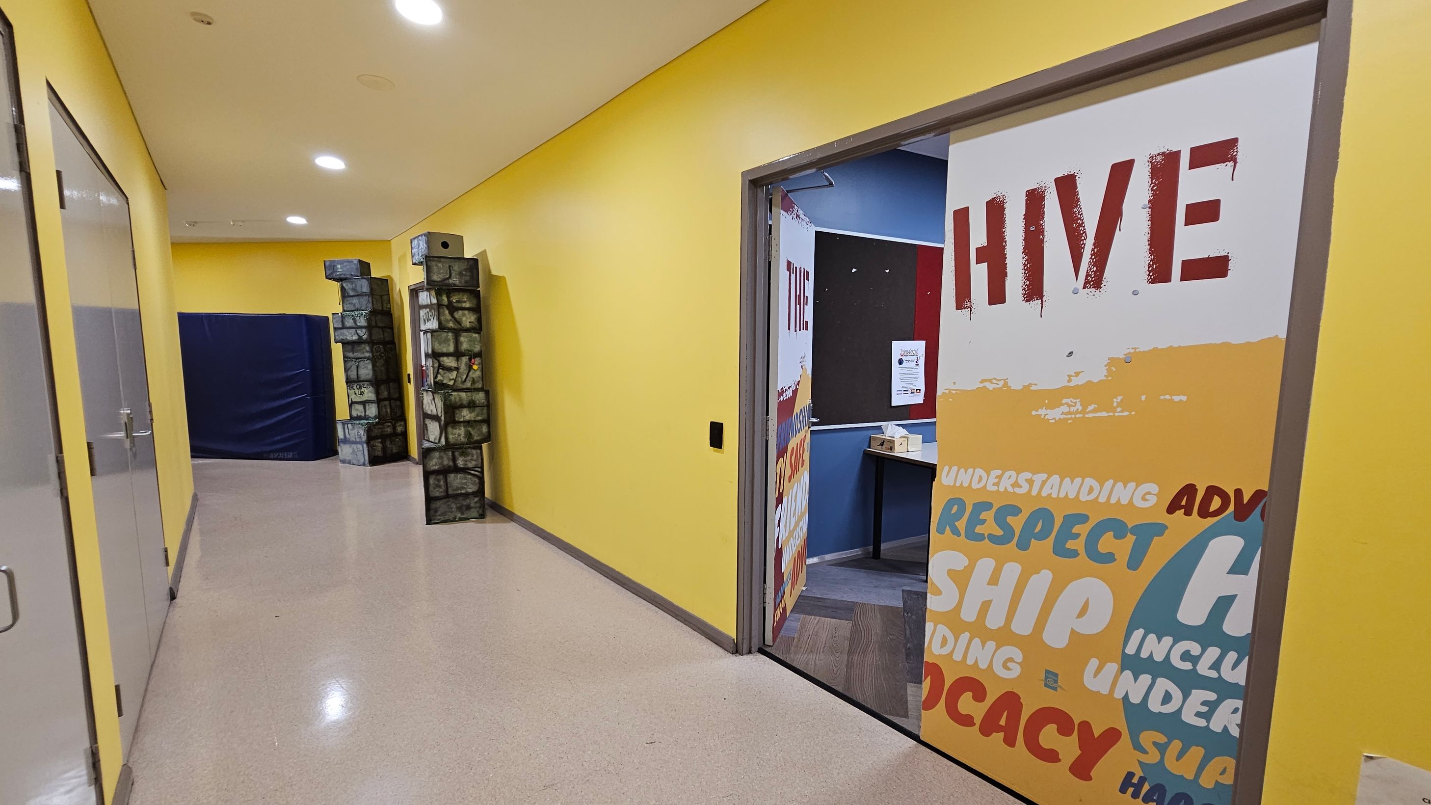 A hallway with yellow walls and a light-coloured floor. On the right, an open door with the word "HIVE" in large red letters and other words like "RESPECT" and "ADVOCACY" in various colours. Inside the room, a bulletin board and a table are visible. On the left, glass doors with metal handles. At the end of the hallway, there are two tall structures resembling stone pillars and a large blue padded mat. The ceiling has recessed lighting. A hallway with yellow walls and a light-coloured floor. On the right, an open door with the word "HIVE" in large red letters and other words like "RESPECT" and "ADVOCACY" in various colours. Inside the room, a bulletin board and a table are visible. On the left, glass doors with metal handles. At the end of the hallway, there are two tall structures resembling stone pillars and a large blue padded mat. The ceiling has recessed lighting.