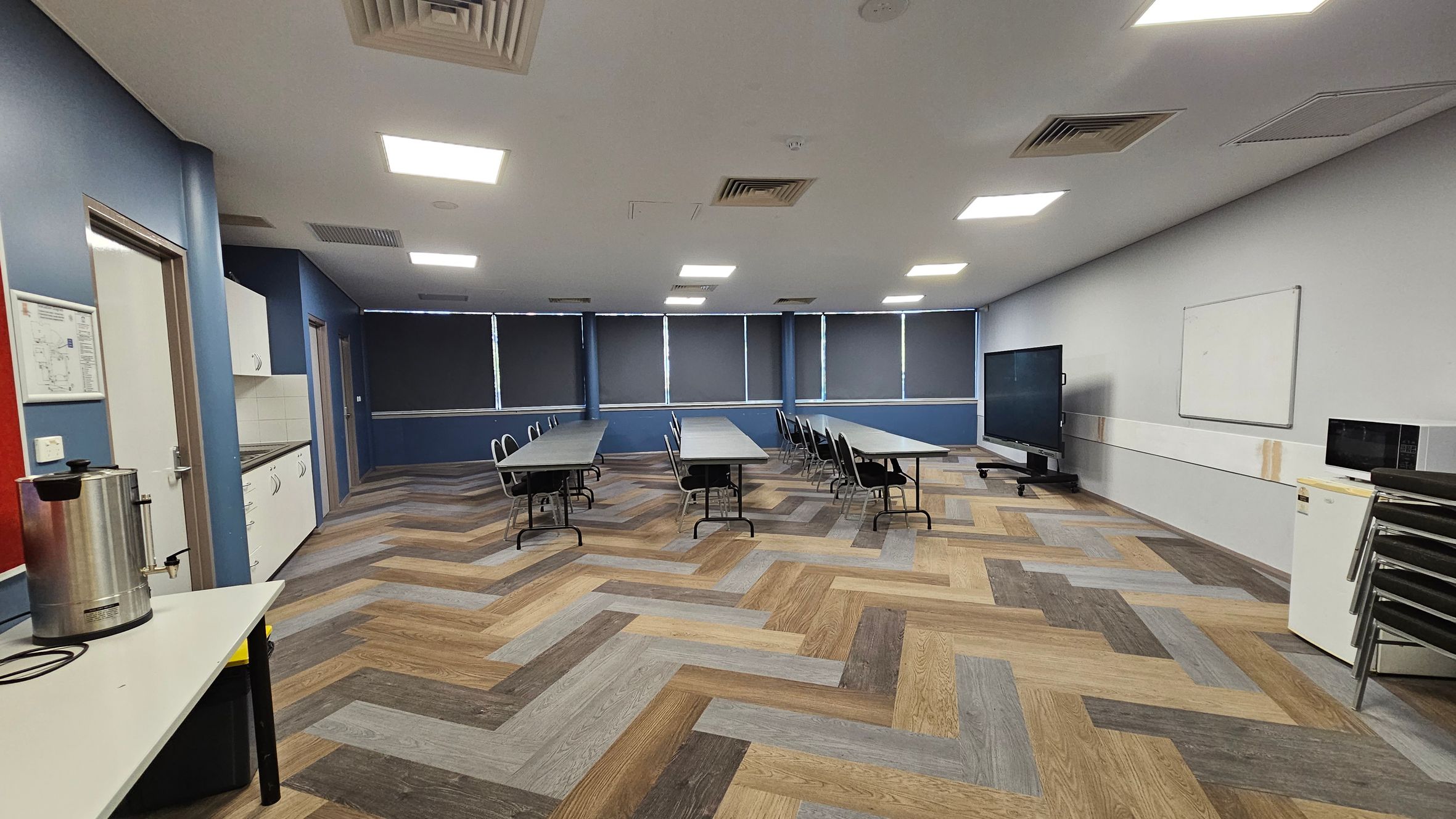 A room with a herringbone-patterned wooden floor and a white ceiling with recessed lights. On the left, there's a counter with a large metal urn and a door. Cabinets and a sink are along the left wall. In the centre, several tables with chairs are arranged in rows. The right side has a large screen on a stand, a whiteboard, and a small fridge with a microwave on top. The windows at the back are covered with dark blinds. A room with a herringbone-patterned wooden floor and a white ceiling with recessed lights. On the left, there's a counter with a large metal urn and a door. Cabinets and a sink are along the left wall. In the centre, several tables with chairs are arranged in rows. The right side has a large screen on a stand, a whiteboard, and a small fridge with a microwave on top. The windows at the back are covered with dark blinds.