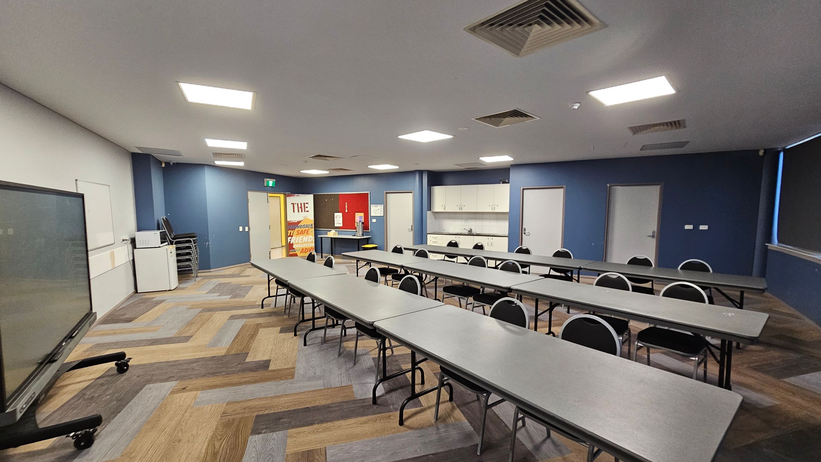 A classroom with a modern design. On the left, there's a large interactive screen on wheels and a whiteboard. The floor has a herringbone pattern with various shades of wood. In the centre, there are long tables with black chairs arranged in rows. The back wall is blue, featuring a door, cabinets, and a sink. On the right, a window with blinds is partially visible. The ceiling has recessed lighting and air vents. A colourful poster is near the back wall. A classroom with a modern design. On the left, there's a large interactive screen on wheels and a whiteboard. The floor has a herringbone pattern with various shades of wood. In the centre, there are long tables with black chairs arranged in rows. The back wall is blue, featuring a door, cabinets, and a sink. On the right, a window with blinds is partially visible. The ceiling has recessed lighting and air vents. A colourful poster is near the back wall.