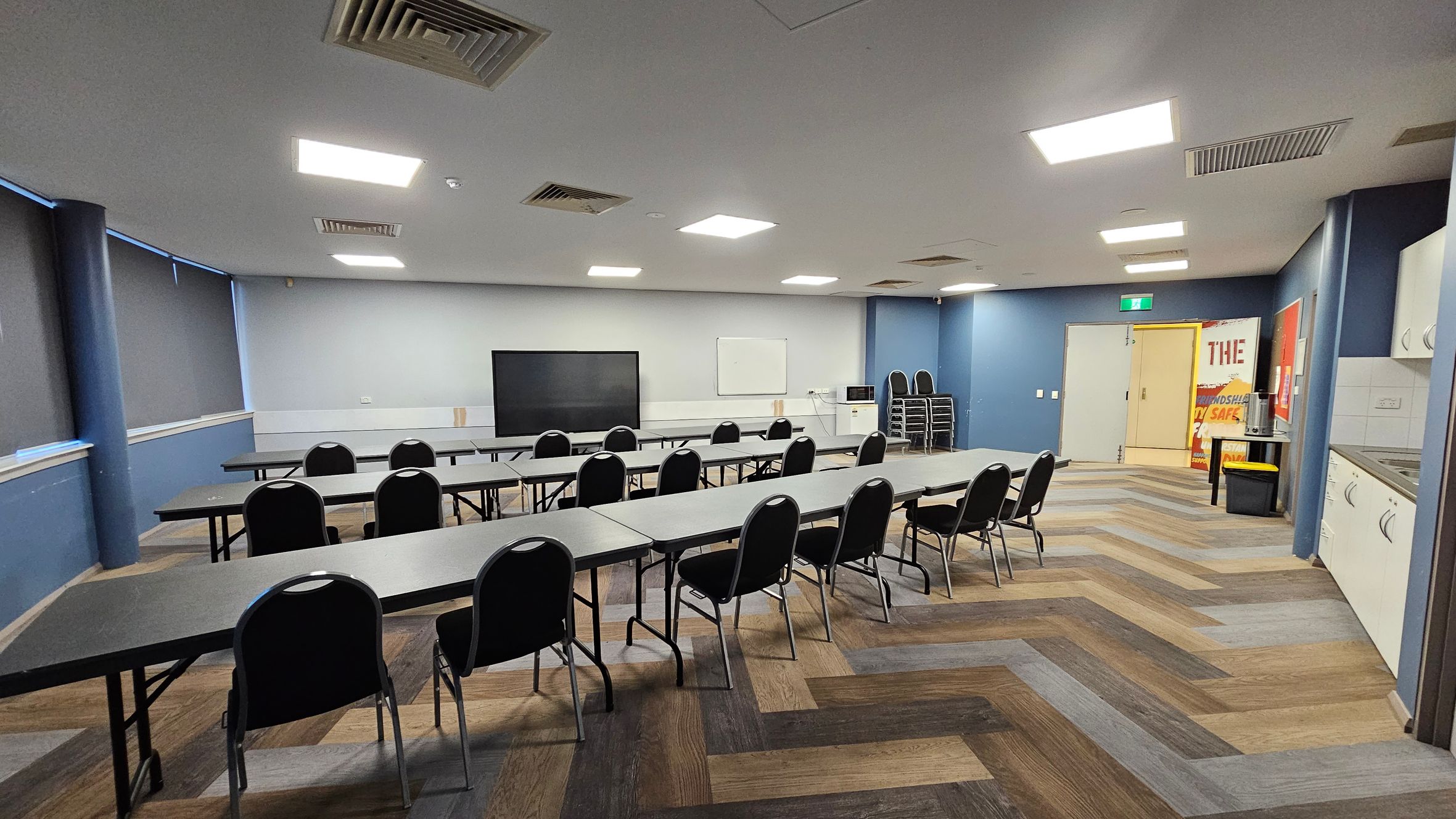 A room with a zigzag patterned wooden floor and a ceiling with recessed lighting and vents. On the left, there are long tables with black chairs facing a large screen and a whiteboard on the wall. The right side has a kitchenette with white cabinets and a stack of chairs. In the background, there's a door with a green exit sign and a poster next to it. The walls are painted blue and grey, and the windows have dark blinds. A room with a zigzag patterned wooden floor and a ceiling with recessed lighting and vents. On the left, there are long tables with black chairs facing a large screen and a whiteboard on the wall. The right side has a kitchenette with white cabinets and a stack of chairs. In the background, there's a door with a green exit sign and a poster next to it. The walls are painted blue and grey, and the windows have dark blinds.