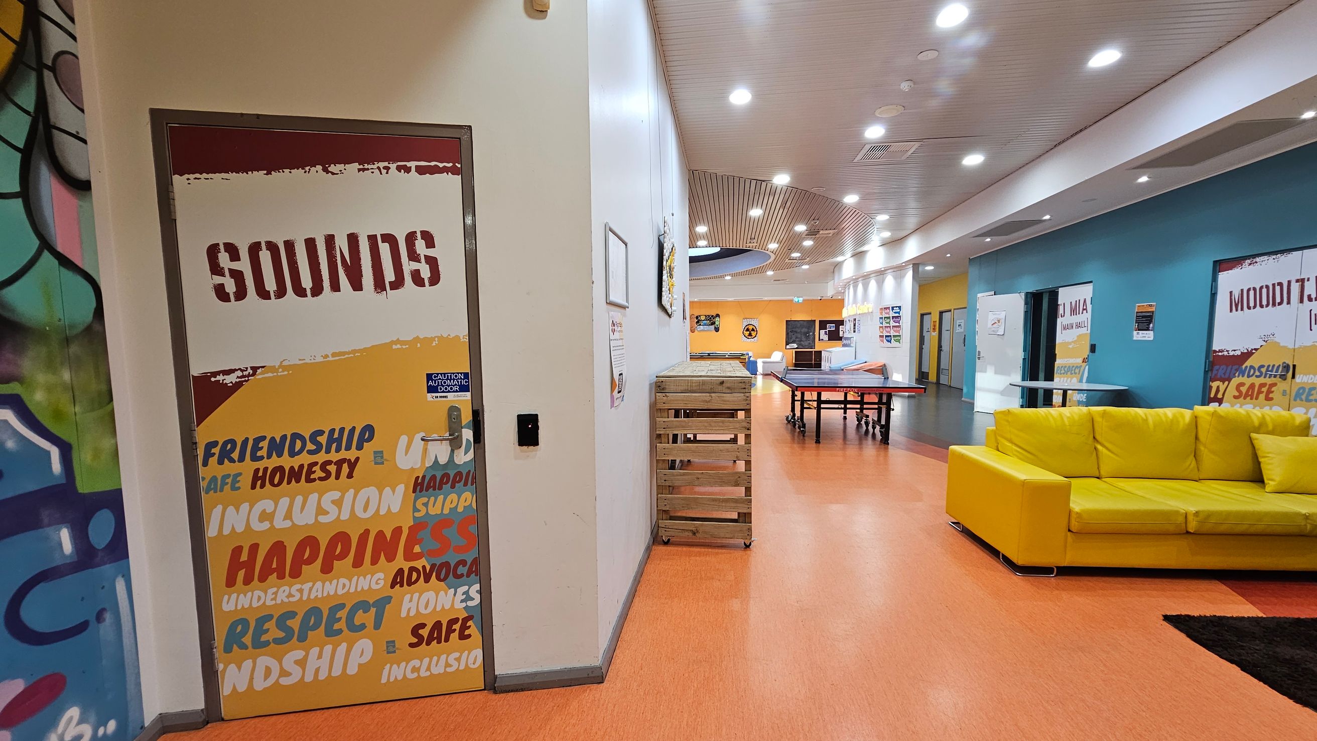 A brightly lit indoor space with an orange floor and a white ceiling featuring recessed lights. On the left, a door labelled "SOUNDS" is decorated with words like "FRIENDSHIP" and "HAPPINESS." Adjacent is a wall with colourful graffiti. In the centre, a wooden crate and a ping pong table are visible. On the right, a vibrant yellow sofa sits against a blue wall with more labelled doors. The corridor extends into the distance, with various posters and signs on the walls. A brightly lit indoor space with an orange floor and a white ceiling featuring recessed lights. On the left, a door labelled "SOUNDS" is decorated with words like "FRIENDSHIP" and "HAPPINESS." Adjacent is a wall with colourful graffiti. In the centre, a wooden crate and a ping pong table are visible. On the right, a vibrant yellow sofa sits against a blue wall with more labelled doors. The corridor extends into the distance, with various posters and signs on the walls.