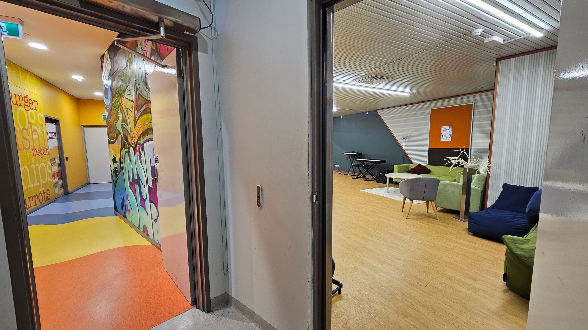 On the left, a hallway with vibrant graffiti art and a yellow wall featuring words like "burger" and "chips." The floor is a mix of blue, yellow, and orange. On the right, a room with a wooden floor, green and blue seating, a grey chair, and a keyboard against a blue wall. The ceiling is lined with white panels and fluorescent lights. On the left, a hallway with vibrant graffiti art and a yellow wall featuring words like "burger" and "chips." The floor is a mix of blue, yellow, and orange. On the right, a room with a wooden floor, green and blue seating, a grey chair, and a keyboard against a blue wall. The ceiling is lined with white panels and fluorescent lights.