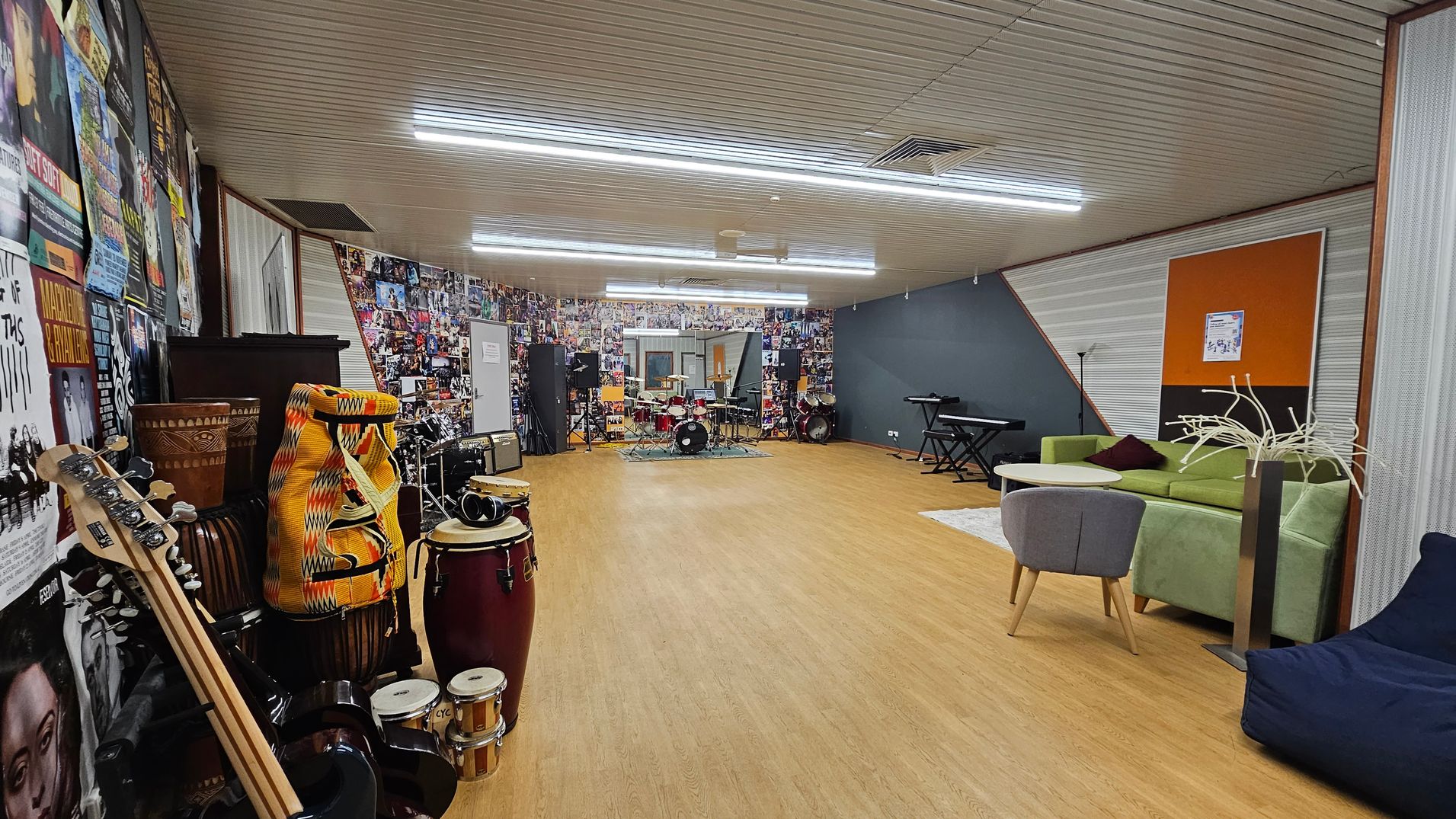 A spacious music room with a wooden floor and a ceiling with fluorescent lights. On the left, there are various musical instruments, including guitars, drums, and percussion instruments, against a wall covered in posters. In the centre, a drum set is visible. On the right, there is a green sofa, a small round table, a grey chair, and a blue bean bag. The back wall is decorated with numerous photos, and a keyboard is positioned nearby. A spacious music room with a wooden floor and a ceiling with fluorescent lights. On the left, there are various musical instruments, including guitars, drums, and percussion instruments, against a wall covered in posters. In the centre, a drum set is visible. On the right, there is a green sofa, a small round table, a grey chair, and a blue bean bag. The back wall is decorated with numerous photos, and a keyboard is positioned nearby.