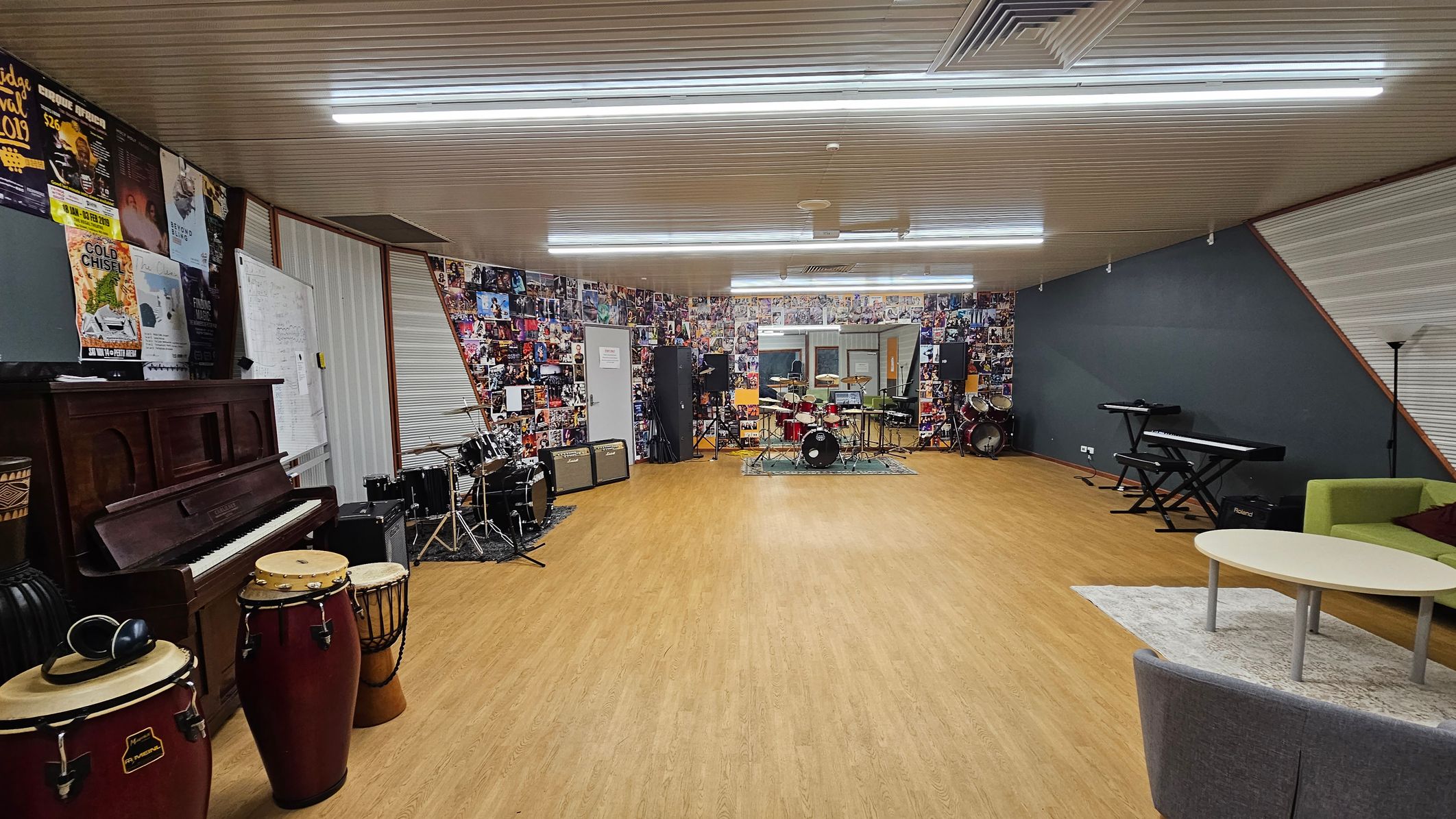A spacious music room with a wooden floor and a ceiling with fluorescent lights. On the left, there is a piano and a set of conga drums. The back wall is covered with posters and features a drum kit and amplifiers. On the right, there is a keyboard on a stand, a green sofa, and a round table on a rug. The walls are decorated with numerous music-related images and posters. A spacious music room with a wooden floor and a ceiling with fluorescent lights. On the left, there is a piano and a set of conga drums. The back wall is covered with posters and features a drum kit and amplifiers. On the right, there is a keyboard on a stand, a green sofa, and a round table on a rug. The walls are decorated with numerous music-related images and posters.