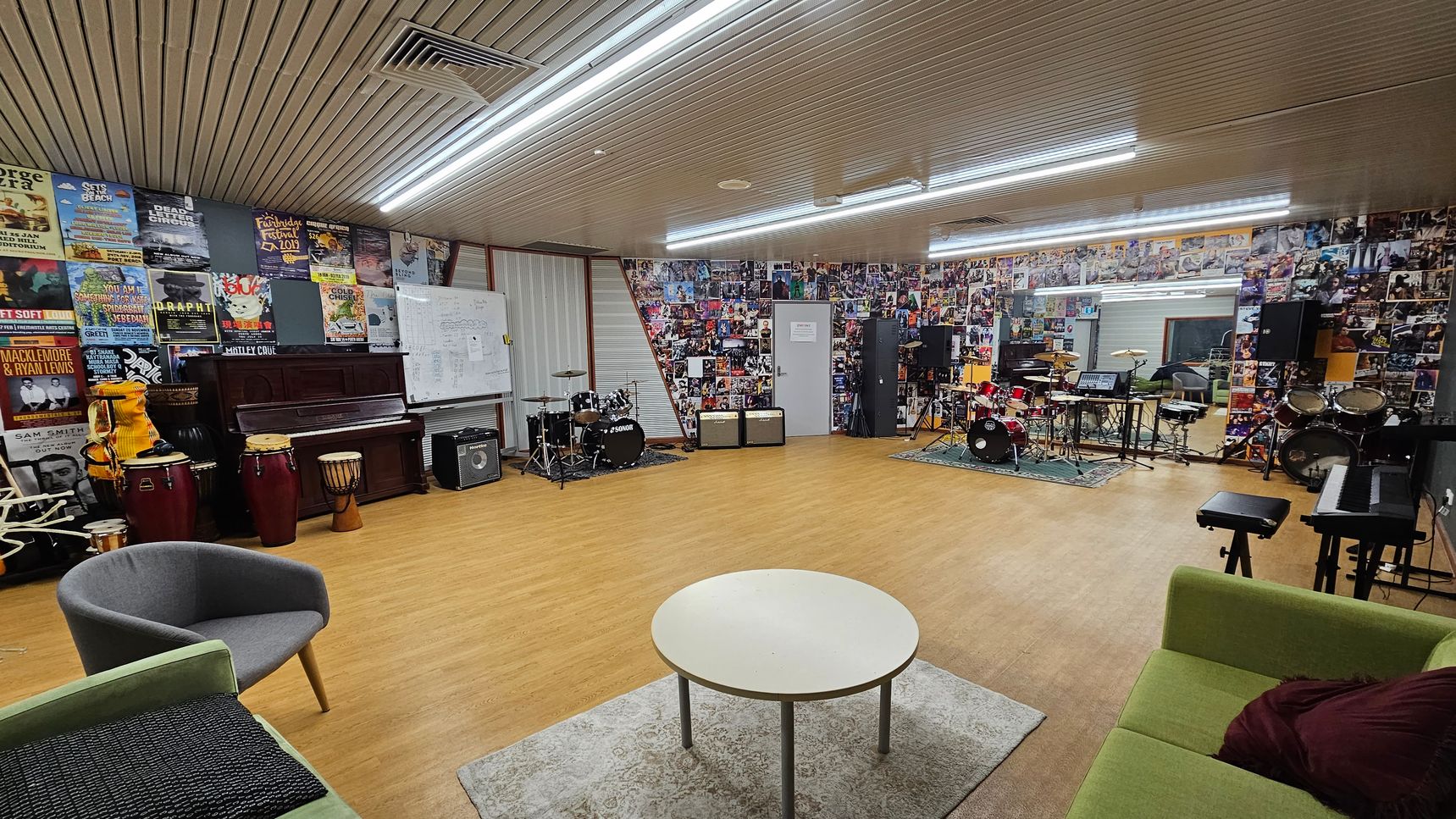 A spacious music room with wooden flooring and a ceiling with fluorescent lights. The left side features a wall covered in posters, a piano, and various percussion instruments. In the centre, there's a round table on a rug, surrounded by chairs and a green sofa. The right side has a wall adorned with photos, multiple drum kits, amplifiers, and a keyboard. A whiteboard is also visible on the left wall. A spacious music room with wooden flooring and a ceiling with fluorescent lights. The left side features a wall covered in posters, a piano, and various percussion instruments. In the centre, there's a round table on a rug, surrounded by chairs and a green sofa. The right side has a wall adorned with photos, multiple drum kits, amplifiers, and a keyboard. A whiteboard is also visible on the left wall.