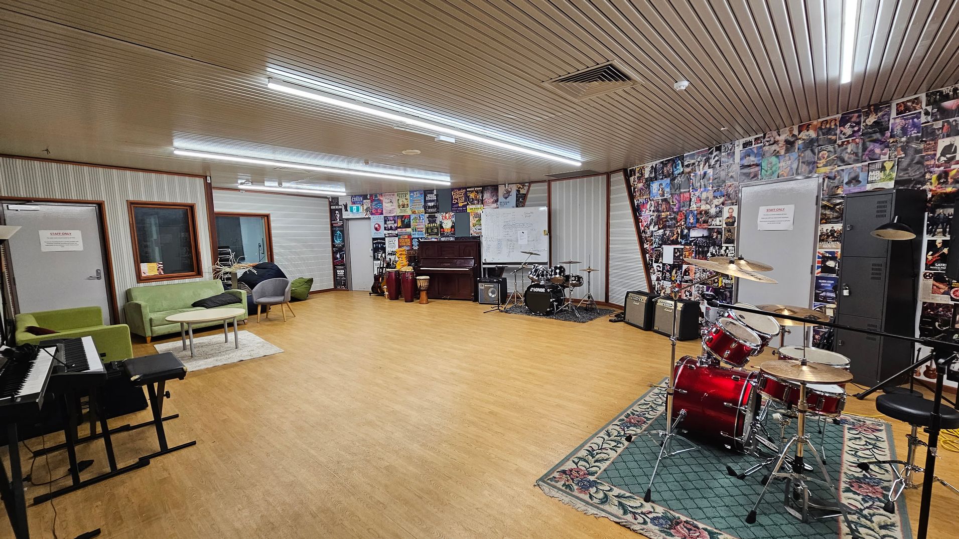 A music room with a wooden floor and a ceiling with fluorescent lights. On the right, a red drum set is placed on a patterned green rug. The back wall is covered with posters and features a piano, conga drums, and amplifiers. A whiteboard is also visible. On the left, there are green sofas and grey chairs around a small table, with a keyboard nearby. The walls have a mix of corrugated and poster-covered surfaces. A music room with a wooden floor and a ceiling with fluorescent lights. On the right, a red drum set is placed on a patterned green rug. The back wall is covered with posters and features a piano, conga drums, and amplifiers. A whiteboard is also visible. On the left, there are green sofas and grey chairs around a small table, with a keyboard nearby. The walls have a mix of corrugated and poster-covered surfaces.