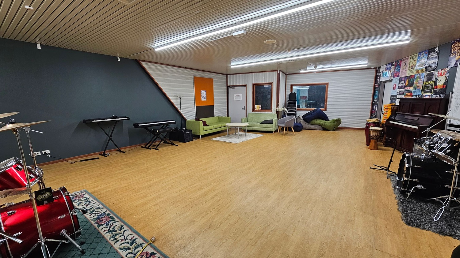 A spacious music room with a wooden floor and a ceiling with fluorescent lights. On the left, there are two electronic keyboards against a dark wall, and a red drum set on a patterned rug. In the centre, green sofas and a small table are placed on a light rug. The right side features a wall with posters, a piano, and additional drums. A window and a door are visible in the background, along with stacked chairs and bean bags. A spacious music room with a wooden floor and a ceiling with fluorescent lights. On the left, there are two electronic keyboards against a dark wall, and a red drum set on a patterned rug. In the centre, green sofas and a small table are placed on a light rug. The right side features a wall with posters, a piano, and additional drums. A window and a door are visible in the background, along with stacked chairs and bean bags.