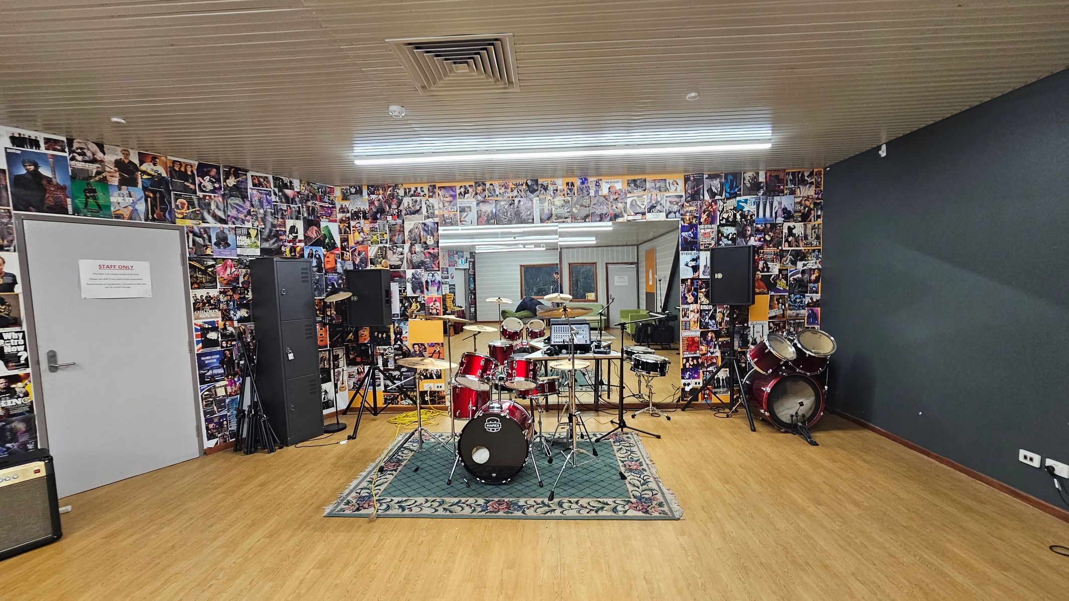 A music rehearsal room with a wooden floor and a ceiling featuring a vent. In the centre, a red drum kit is set on a patterned rug. The back wall is covered with a collage of music posters and a large mirror. To the left, a grey door marked "Staff Only" is visible, alongside speakers and equipment. On the right, additional drums and speakers are present. The room is well-lit with fluorescent lights. A music rehearsal room with a wooden floor and a ceiling featuring a vent. In the centre, a red drum kit is set on a patterned rug. The back wall is covered with a collage of music posters and a large mirror. To the left, a grey door marked "Staff Only" is visible, alongside speakers and equipment. On the right, additional drums and speakers are present. The room is well-lit with fluorescent lights.