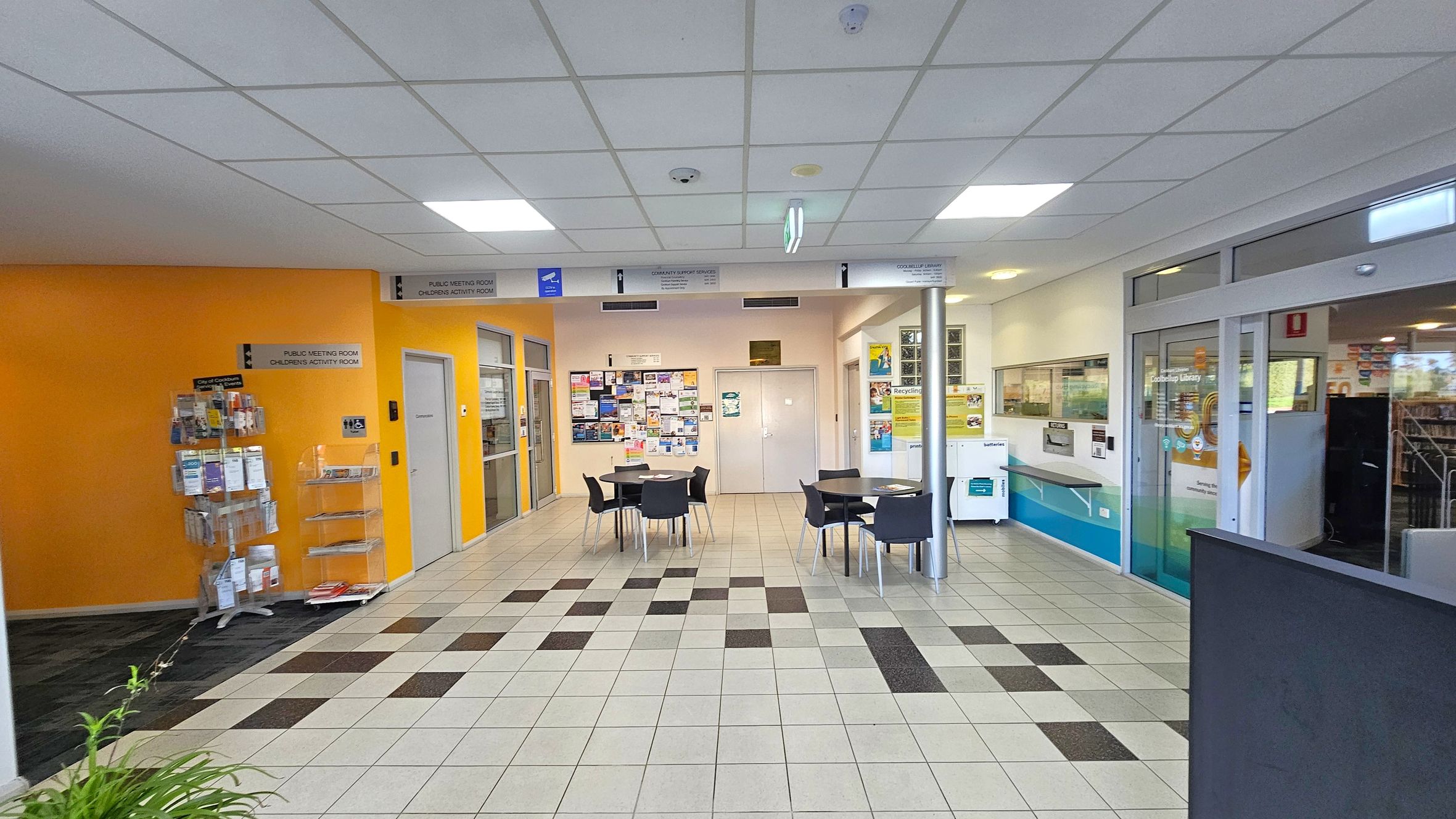 A library entrance area with a tiled floor featuring a pattern of light and dark squares. On the left, there's a bright orange wall with a brochure stand. In the centre, several round tables with black chairs are arranged. The back wall has a bulletin board filled with notices. On the right, glass doors lead into the library, with a sign above them. The ceiling is white with recessed lighting.