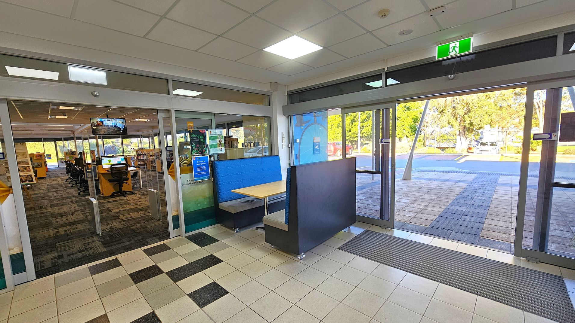 A library entrance with glass doors on the left leading to an interior with bookshelves and computer stations. A blue booth with a wooden table is positioned near the entrance. On the right, large glass doors open to an outdoor area with trees and a parked car visible. The floor is tiled, and the ceiling has recessed lighting and an illuminated exit sign.