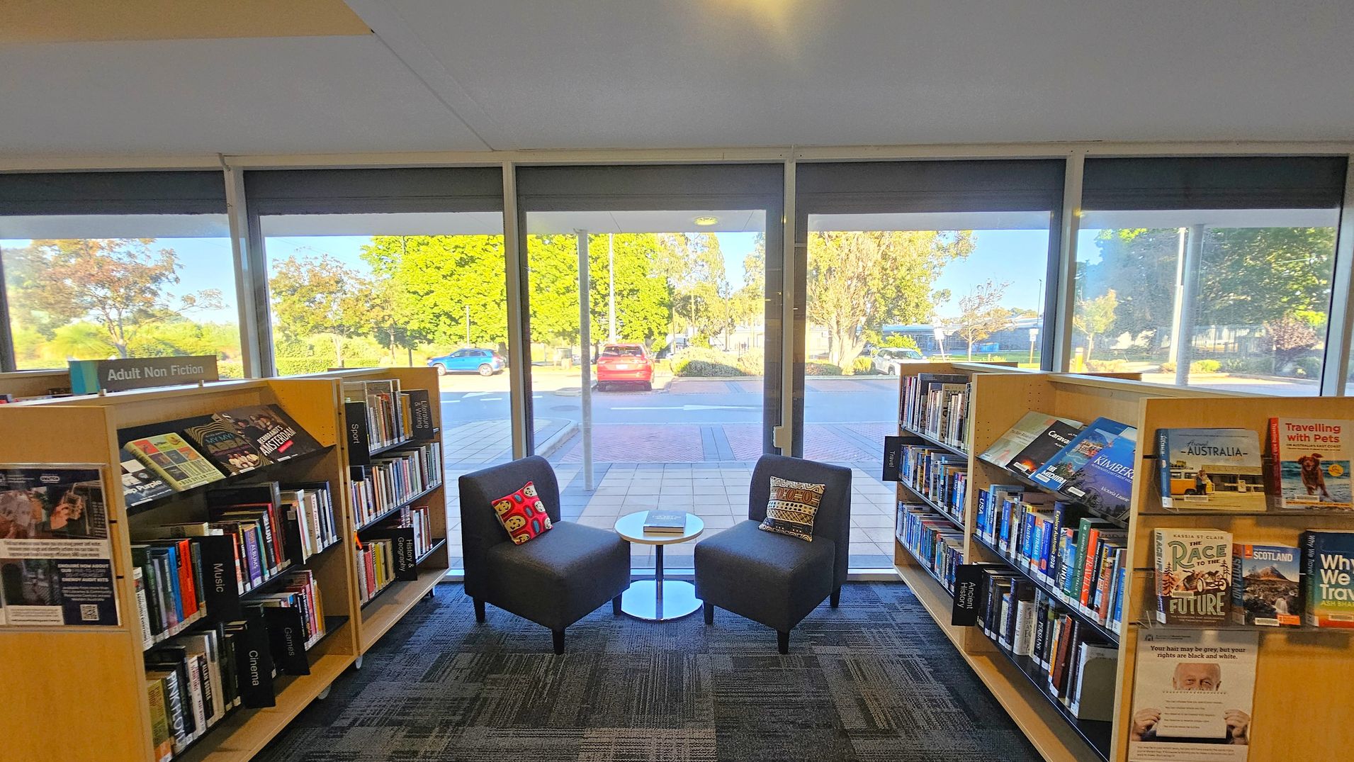 Bookshelves filled with books line the left and right sides, labelled "Adult Non Fiction." In the centre, two grey chairs with patterned cushions face each other, separated by a small round table with a book on it. Large windows in the background reveal a view of parked cars and greenery outside. The floor is carpeted with a geometric pattern, and the ceiling is plain with a light fixture.