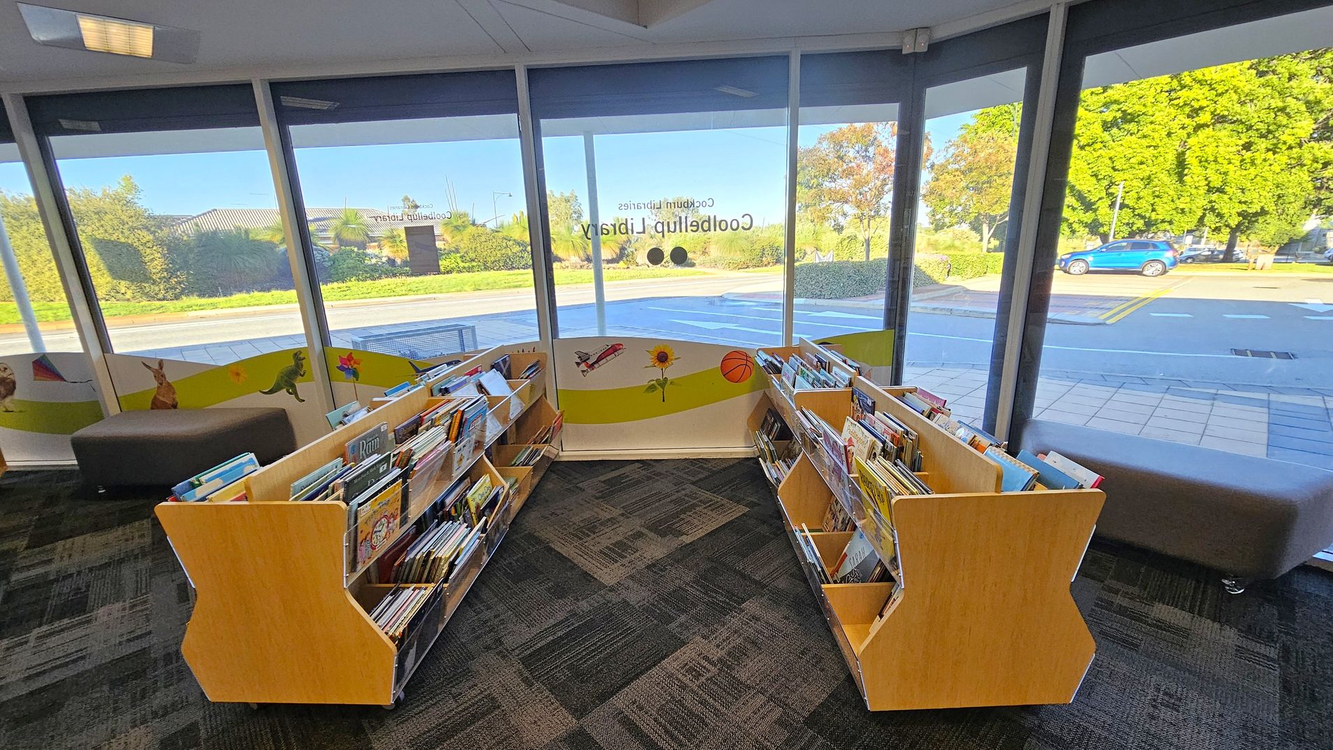 Inside a library, two wooden bookshelves filled with children's books are positioned in a V-shape. The floor is carpeted with a geometric pattern. Large windows in the background provide a view of the outside, showing a road, greenery, and a blue car. The windows have colourful decals featuring a kangaroo, dinosaur, aeroplane, sunflower, and basketball. Grey cushioned seating is visible along the windows.