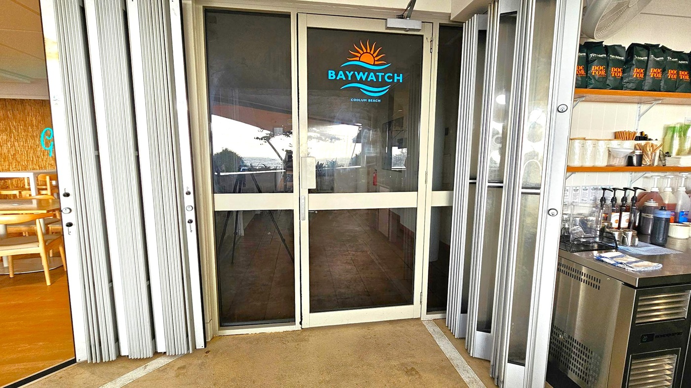 Glass door with "Baywatch Coolum Beach" logo in the centre, featuring a sun and wave design. On the left, a partially visible dining area with wooden chairs and tables. On the right, a counter with coffee supplies, including cups, syrups, and a coffee machine. The floor is light-coloured and slightly textured. The ceiling is not visible.