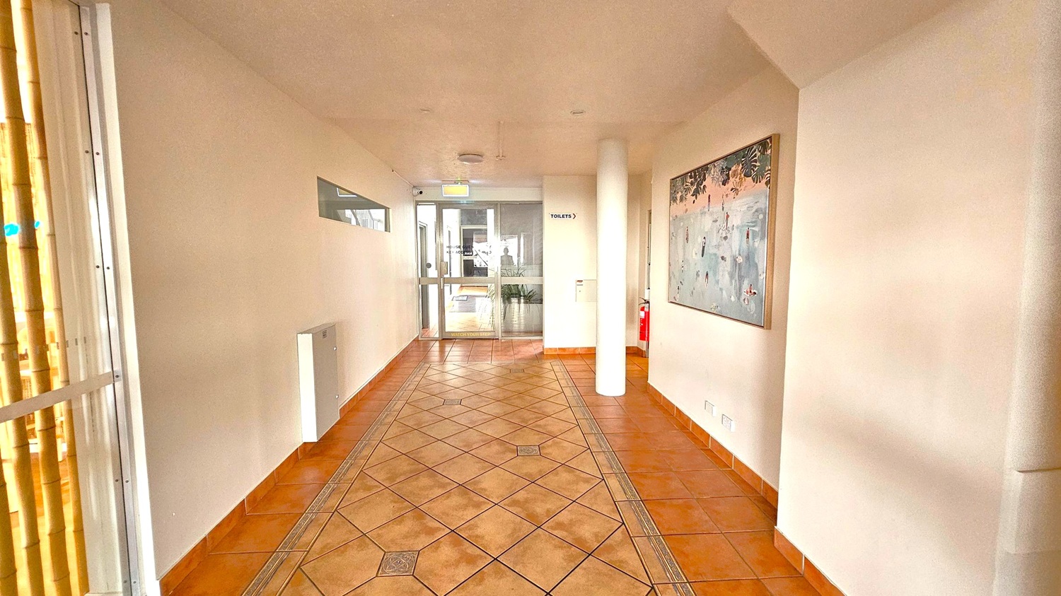 A corridor with terracotta-coloured tiled flooring featuring a diamond pattern. On the right wall, there's a large painting with abstract designs. A white column stands near the painting. The left side has a window with vertical blinds. At the end of the corridor, there's a glass door leading to another area, with a sign indicating toilets to the right. A fire extinguisher is mounted on the right wall near the door. The ceiling is plain and light-coloured.