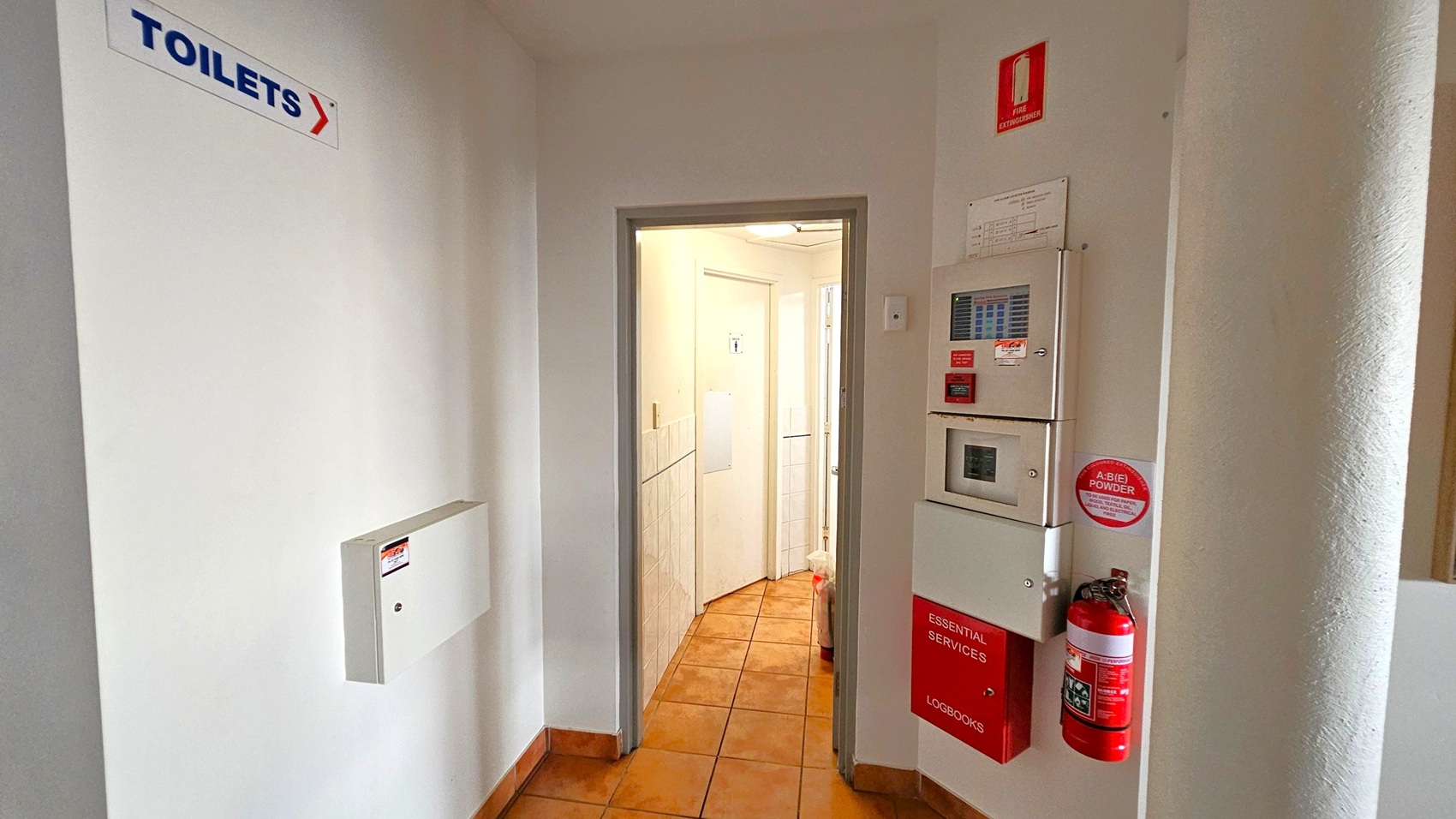 A hallway with a sign on the left wall indicating the direction to the toilets. The floor is covered with brown tiles. On the right wall, there is a fire extinguisher, a fire alarm panel, and a box labelled "Essential Services Logbooks." The walls are painted white, and there is a doorway leading to a well-lit area with more doors visible in the background.