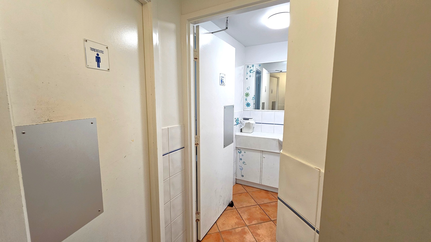 A bathroom entrance with a partially open door on the left, displaying a sign with a figure. Inside, a white sink with a paper towel dispenser is visible, set against a wall with floral tile patterns. The floor is covered with terracotta tiles, and a round ceiling light illuminates the space.