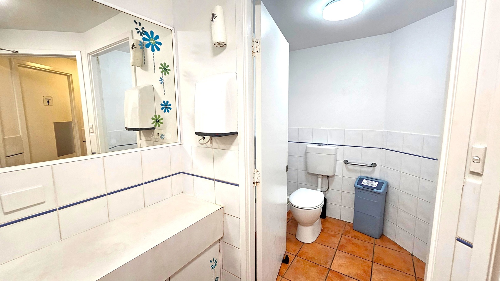 A bathroom with a white tiled wall and terracotta floor tiles. On the left, a large mirror with floral decorations reflects the room. Below the mirror, a beige counter is visible. A white paper towel dispenser is mounted on the wall. On the right, a white toilet with a cistern is next to a grey bin. A grab bar is installed on the wall beside the toilet. The ceiling has a round light fixture.