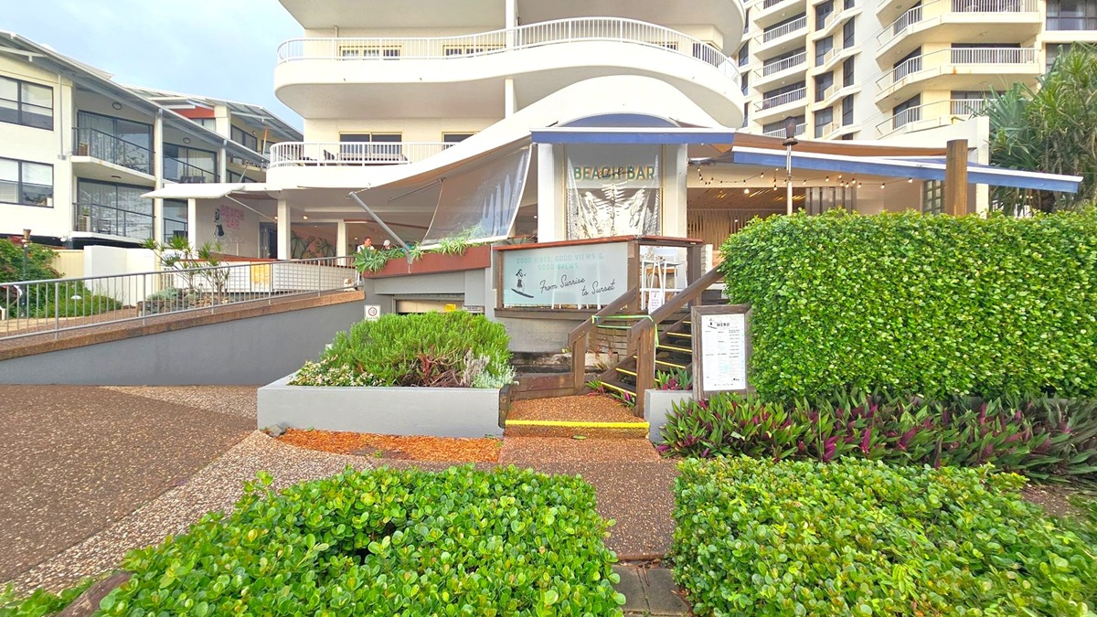 A beach bar with a modern design is in the centre, featuring a curved roof and a sign that reads "BEACH BAR." A menu board is visible on the right, next to a staircase leading up to the entrance. Lush green hedges and plants surround the area, with a paved path in the foreground. The background shows a multi-storey building with balconies, and another building with large windows on the left.