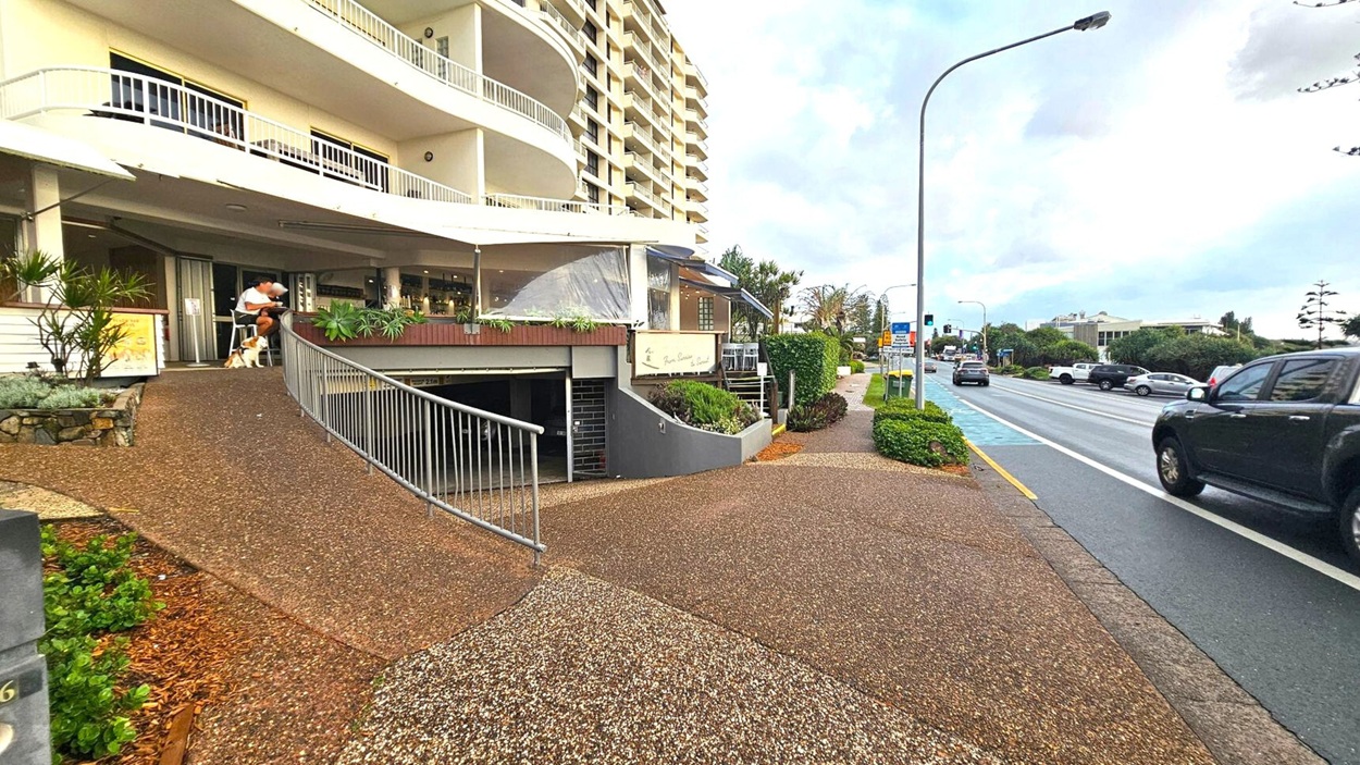 A street scene with a multi-storey building on the left featuring balconies and a café with outdoor seating. A person is sitting at a table with a dog nearby. A sloped path with a railing leads downwards. On the right, a road with several vehicles, including a black SUV, runs parallel to the building. The pavement is bordered by greenery, and a streetlight is visible. The sky is overcast, and the area appears to be a mix of residential and commercial spaces.