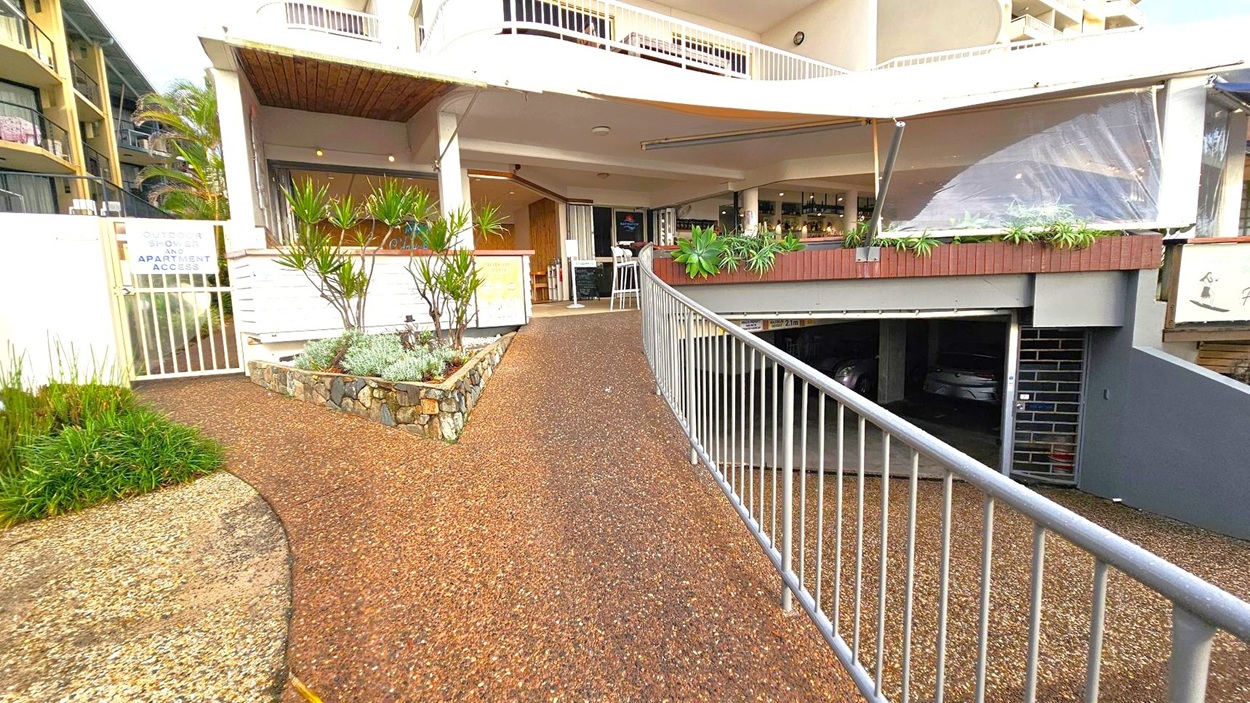 A pathway with a textured surface leads towards a building entrance. On the left, there's a gate with a sign for outdoor shower and apartment access, surrounded by plants and a stone border. On the right, a railing runs alongside a ramp leading to an underground car park. Above, a balcony with a white railing and a covered area with plants is visible. The building has a modern design with a mix of materials and greenery.