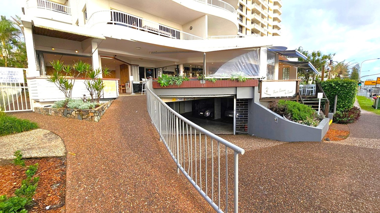 A building with a curved design features a ramp leading to an underground car park on the right. Above the car park entrance, there is a small garden with potted plants. To the left, a stone planter with greenery is adjacent to a sign indicating outdoor shower and apartment access. The building has balconies with white railings. A pathway with a textured surface runs alongside the structure, and there are tables and chairs visible near the entrance.