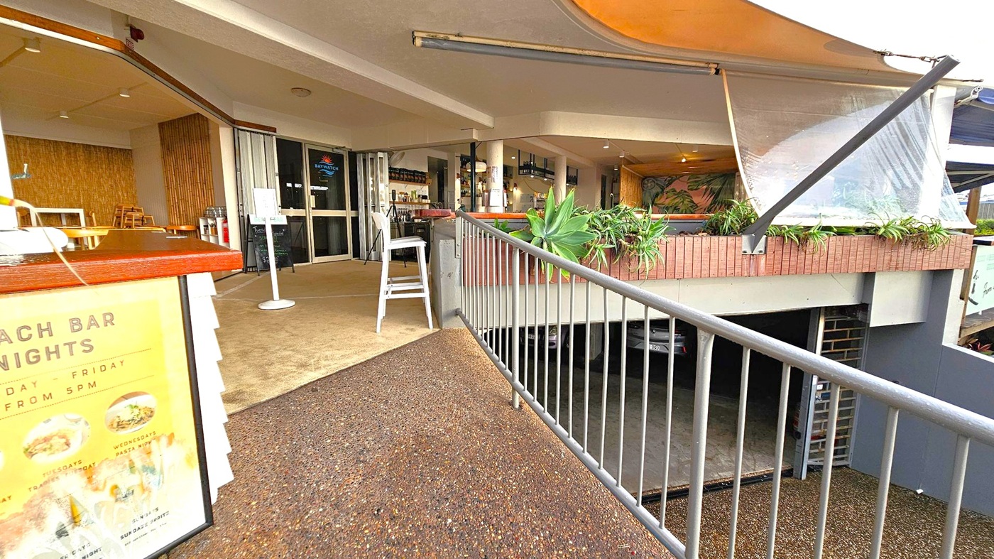 A covered outdoor area with a textured floor and a metal railing leading to a bar entrance. On the left, there's a sign with promotional information. The entrance features glass doors and a sign above them. Inside, there are tables, chairs, and a bar counter with bottles on shelves. Potted plants are placed along the railing and on the brick ledge. The ceiling is flat with recessed lighting, and a canopy extends overhead. Below, a parking area is partially visible.