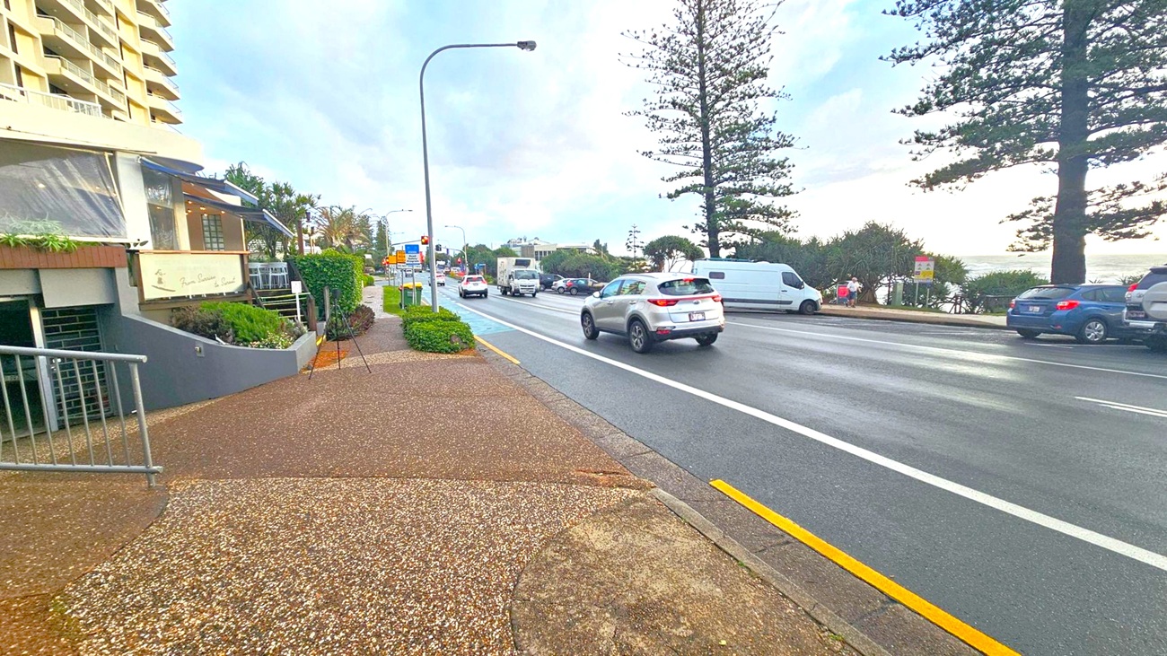 On the left, a building with balconies and a sign is visible, with a small garden area in front. The pavement is textured and leads to a railing. In the centre, a road with several cars, including a white van and a silver SUV, is bordered by a yellow line. On the right, large trees and parked cars line the street, with a glimpse of the ocean in the background. The sky is overcast, and the road appears wet.