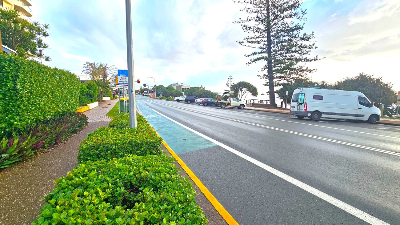 A road with a cycle lane on the left, bordered by lush green hedges and a footpath. Several signs are visible along the path. On the right, a white van is parked by the roadside, with a row of trees and bushes behind it. Further down the road, a few more vehicles are parked. The background features a building and more trees, with a cloudy sky above.