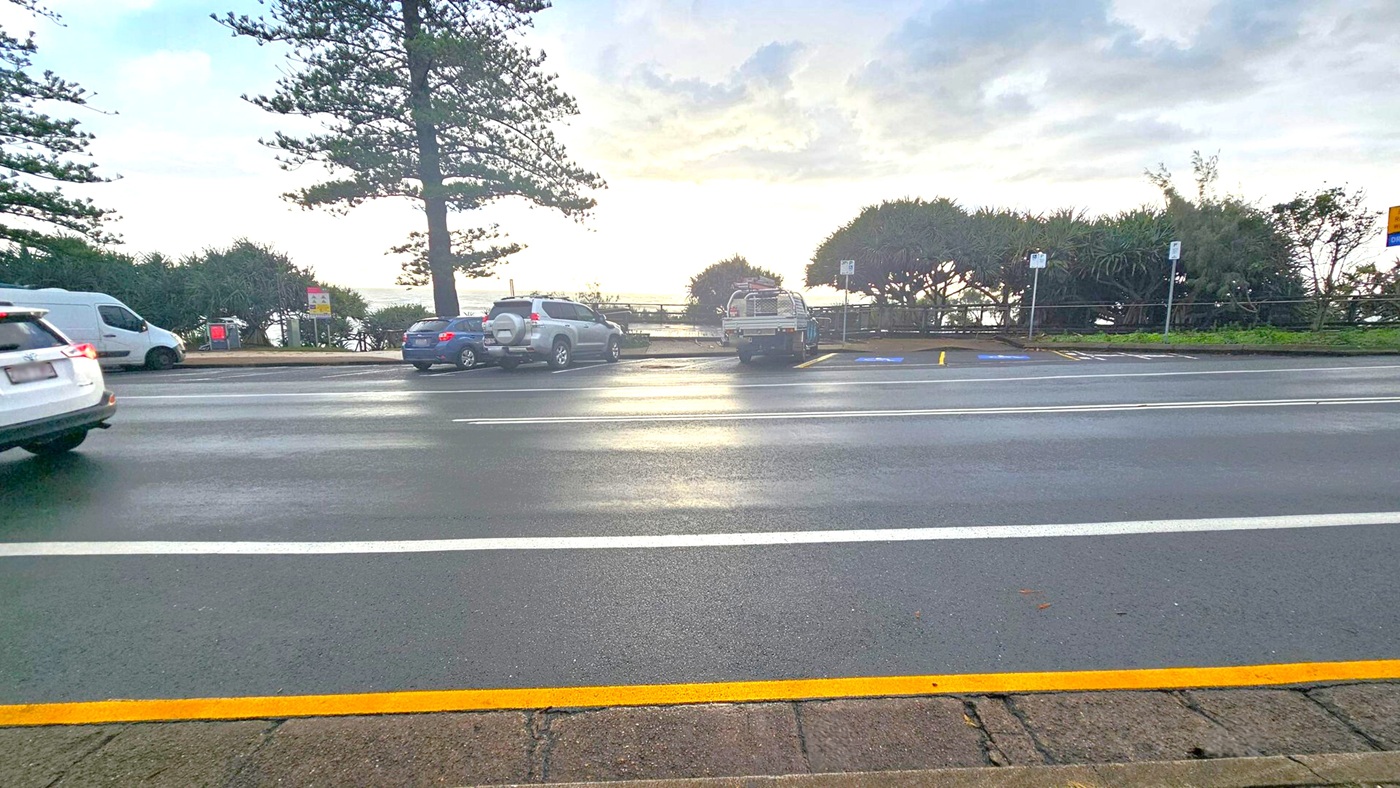A road with a yellow line in the foreground, leading to a parking area with several vehicles, including a white van and a blue car. Tall trees and dense bushes are in the background, with a glimpse of the ocean visible through the foliage. The sky is overcast, and the road surface appears wet, suggesting recent rain.