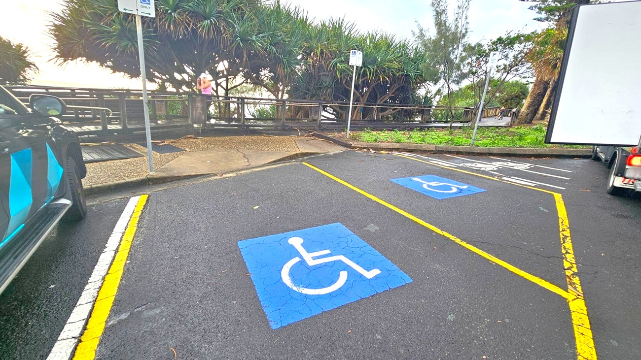 Two parking spaces with blue wheelchair symbols on the asphalt, outlined by yellow lines. On the left, part of a black vehicle is visible. On the right, the rear of a white truck is seen. In the background, a wooden boardwalk with railings is surrounded by dense greenery and trees. A person stands on the boardwalk, partially obscured by foliage. A signpost is present near the boardwalk. The ground is paved, and the area appears to be outdoors.