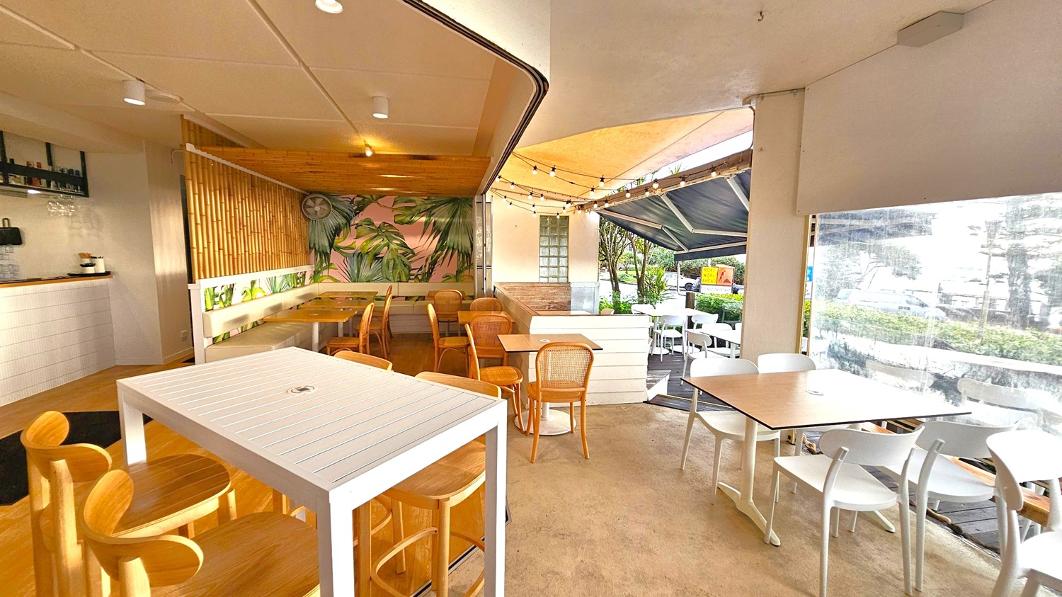 A café interior with a mix of indoor and outdoor seating. On the left, there is a bar area with a white tiled counter and wooden stools. The middle features a bamboo wall with a mural of green leaves and a fan above a bench with wooden chairs. On the right, there are tables with white and wooden chairs, leading to an outdoor area with string lights and a view of greenery and parked cars. The ceiling is white with recessed lighting, and the floor is a light brown.