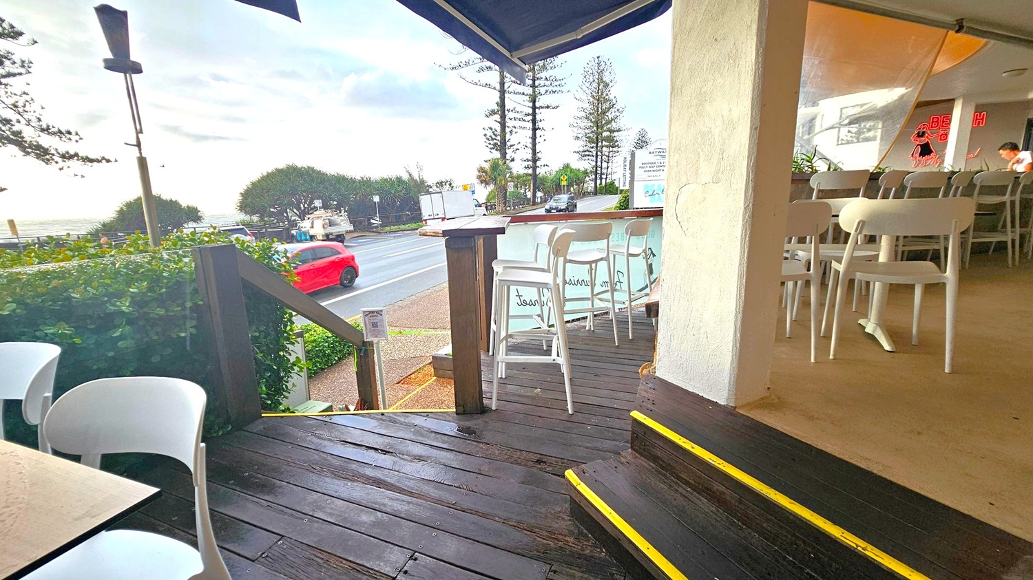 A café setting with a wooden deck in the foreground, featuring white chairs and tables. On the right, the interior has more seating and a neon sign. A red car and a white vehicle are visible on the road outside. The background shows trees and the ocean under a cloudy sky. The floor is wooden with yellow-edged steps, and the ceiling is not visible.