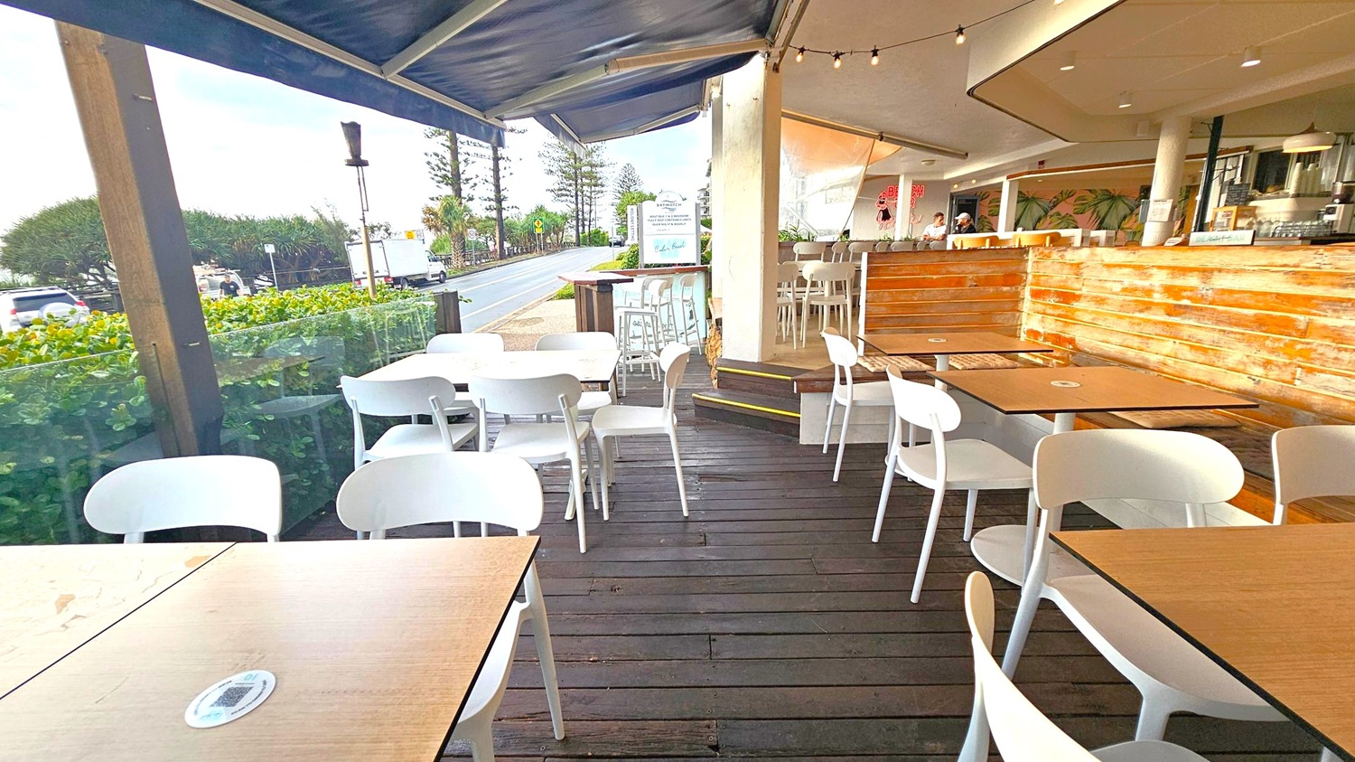 Outdoor café seating area with wooden flooring and white tables and chairs. On the right, a wooden panelled bar with high stools. The ceiling has string lights. In the background, a road lined with trees and a few vehicles. A glass barrier separates the seating area from the greenery outside.
