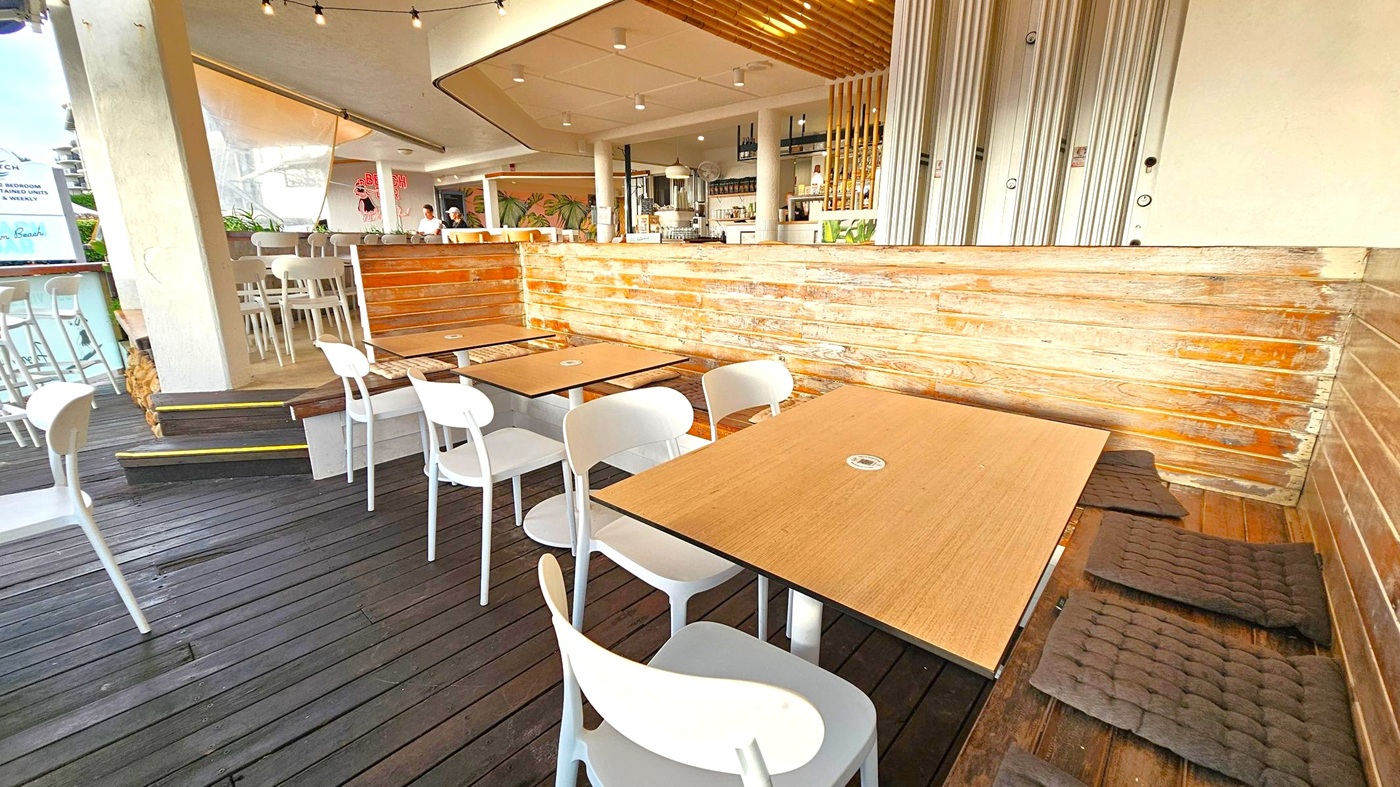 Outdoor café with wooden flooring and light wooden tables surrounded by white chairs. Cushions are placed on the floor near the tables. A wooden panelled wall separates the seating area from the bar, which is visible in the background. The bar area has high stools and a variety of bottles on shelves. Ceiling lights and string lights are visible above. A sign and some greenery are seen on the left side.
