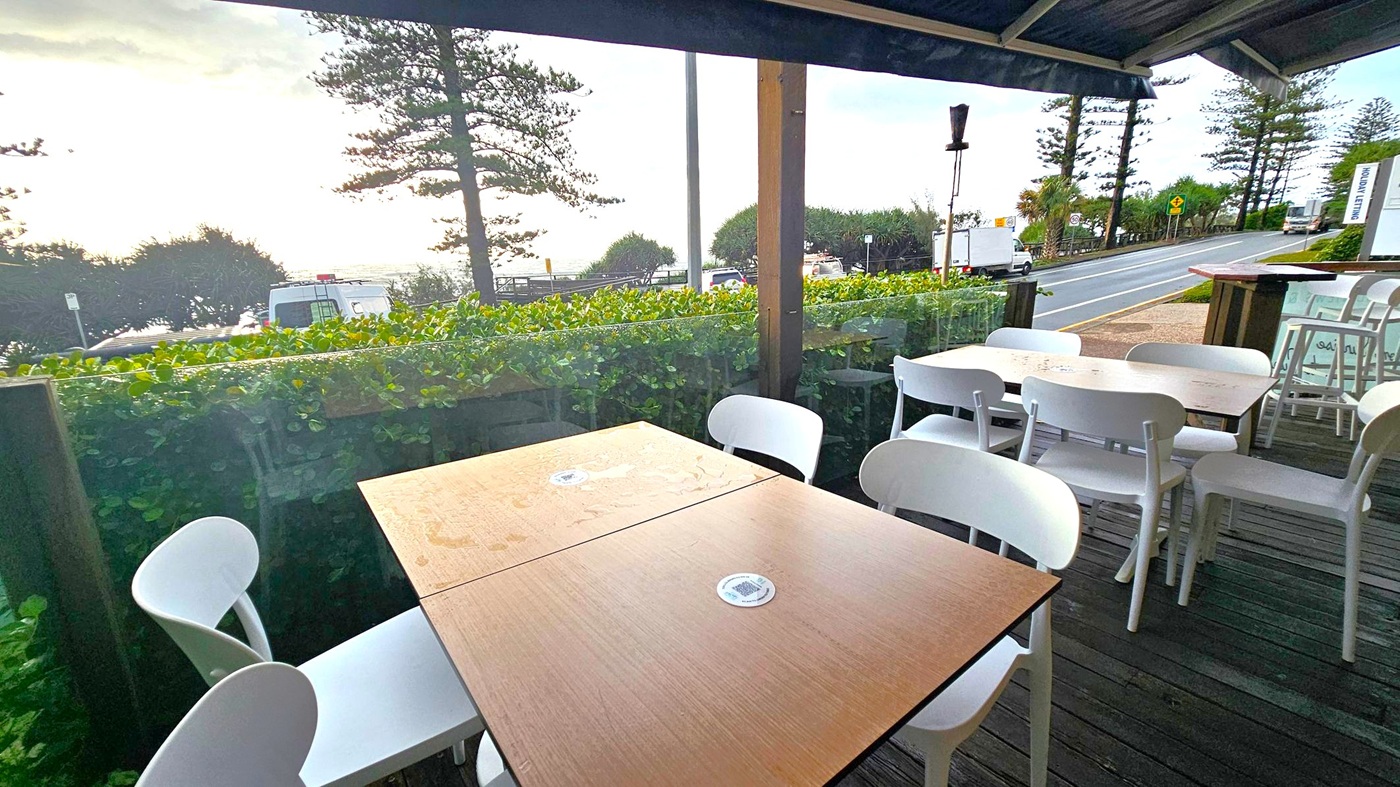 Outdoor seating area with wooden tables and white chairs on a wooden deck. In the background, a road runs parallel to the seating area, with a few vehicles visible. Beyond the road, there are green bushes and tall trees, with the ocean visible in the distance. The sky is overcast, and the area is covered by a roof.