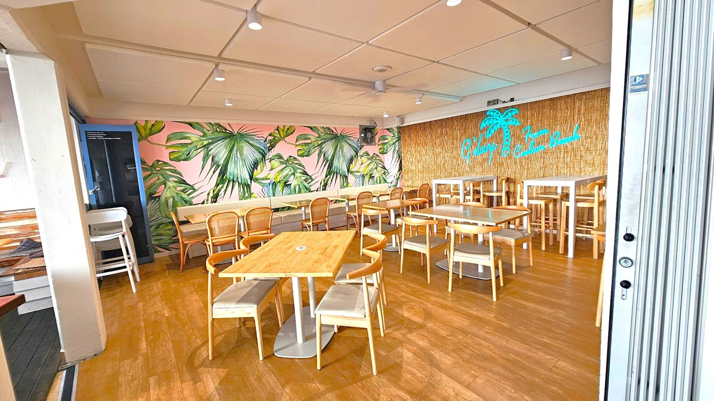 A café interior with wooden flooring and a white panelled ceiling. On the left, there are wooden tables and chairs with a mural of large green leaves on a pink background. The right side features a wall with a straw-like texture and a neon sign of a palm tree and text. High tables and stools are positioned against this wall. The space is well-lit with ceiling lights, and a stack of high chairs is visible in the corner.