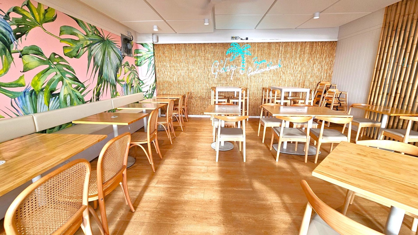 A café interior with wooden flooring and a ceiling with recessed lights. On the left, a wall features a large mural of green tropical leaves on a pink background, accompanied by a beige cushioned bench and wooden tables with rattan chairs. The right side has a bamboo wall with a neon sign of a palm tree and text. Several wooden tables and chairs are arranged neatly, with some stacked in the back.