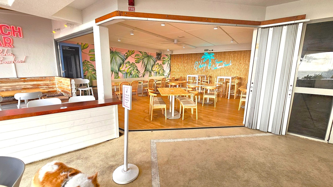 A café interior with a tropical theme. On the left, there's a seating area with white chairs and a wooden bench against a wall with neon signage. The middle features wooden tables and chairs on a wooden floor, with a wall decorated with large leaf patterns. A neon sign with palm tree imagery is on the back wall. On the right, a folding door is partially open. The foreground has a beige carpeted floor and a signpost. A dog is partially visible at the bottom.