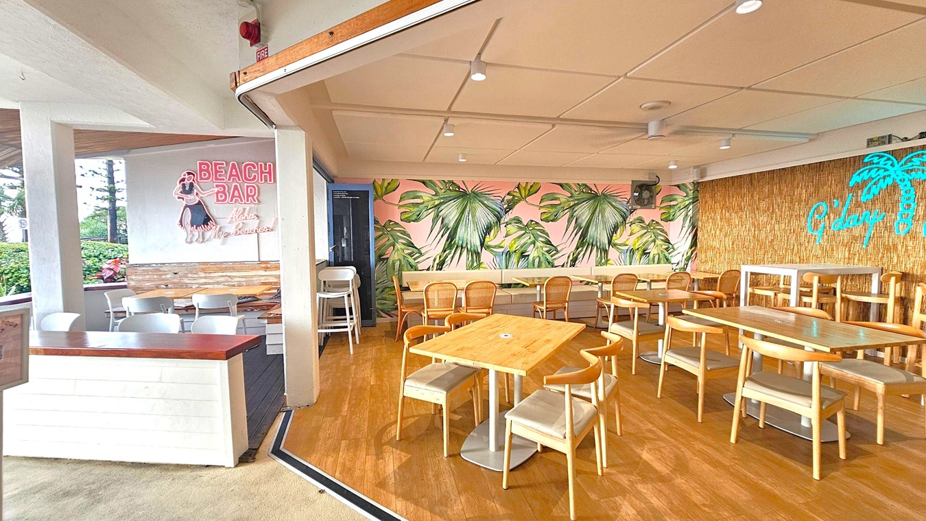 A beach-themed bar interior with a tropical vibe. On the left, a neon sign reads "BEACH BAR" with an illustration of a hula dancer. Below it, white chairs and a wooden table are arranged. The right side features a wall with a large palm leaf mural and a neon sign saying "G'day" next to a palm tree. Wooden tables and chairs fill the space, and the floor is wooden. The ceiling is beige with recessed lighting.