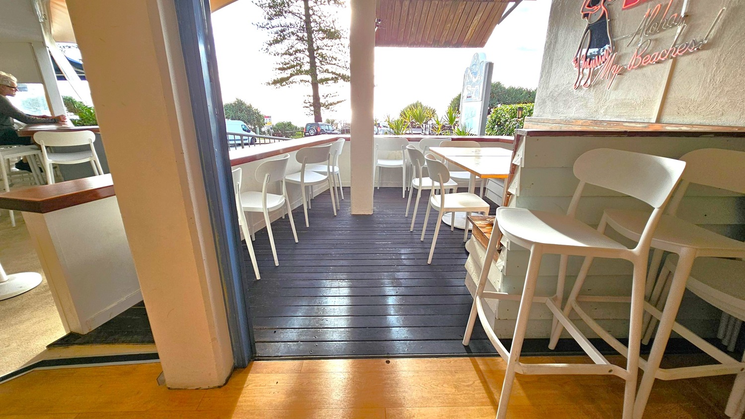 A small outdoor seating area with white chairs and wooden tables on a dark wooden deck. On the left, a person is seated at a table near a window. The right side features a wall with a neon sign and stacked chairs. In the background, there are trees, a parked car, and a glimpse of the ocean. The ceiling is wooden, and the floor inside is light-coloured wood.