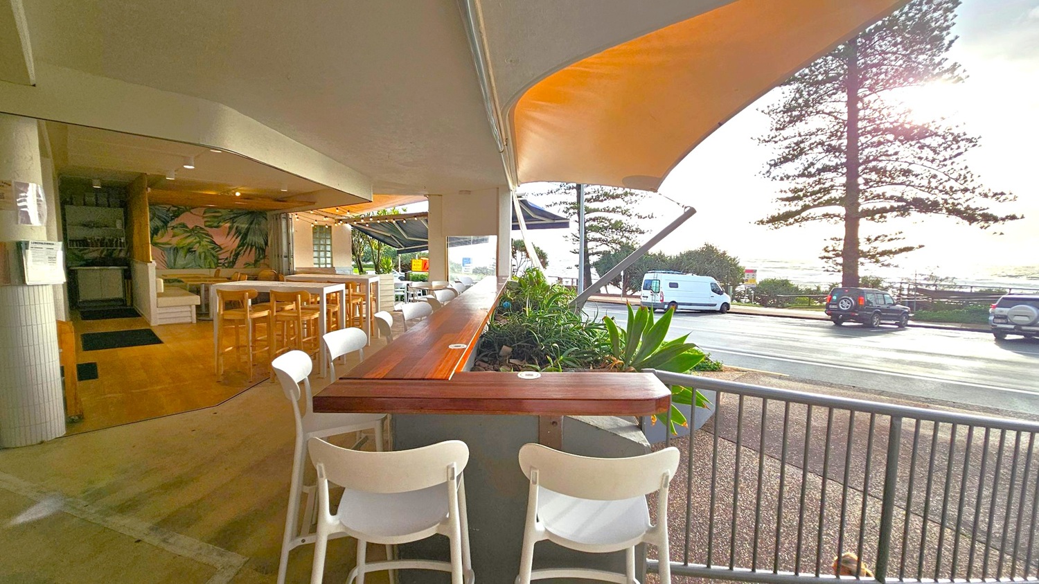 An outdoor café with wooden tables and white chairs on the left, featuring a tropical-themed wall. The seating area is partially covered by a canopy. In the foreground, a wooden counter with plants separates the café from the street. On the right, a road with parked vehicles and a large tree is visible. Beyond the road, the ocean can be seen under a bright sky.